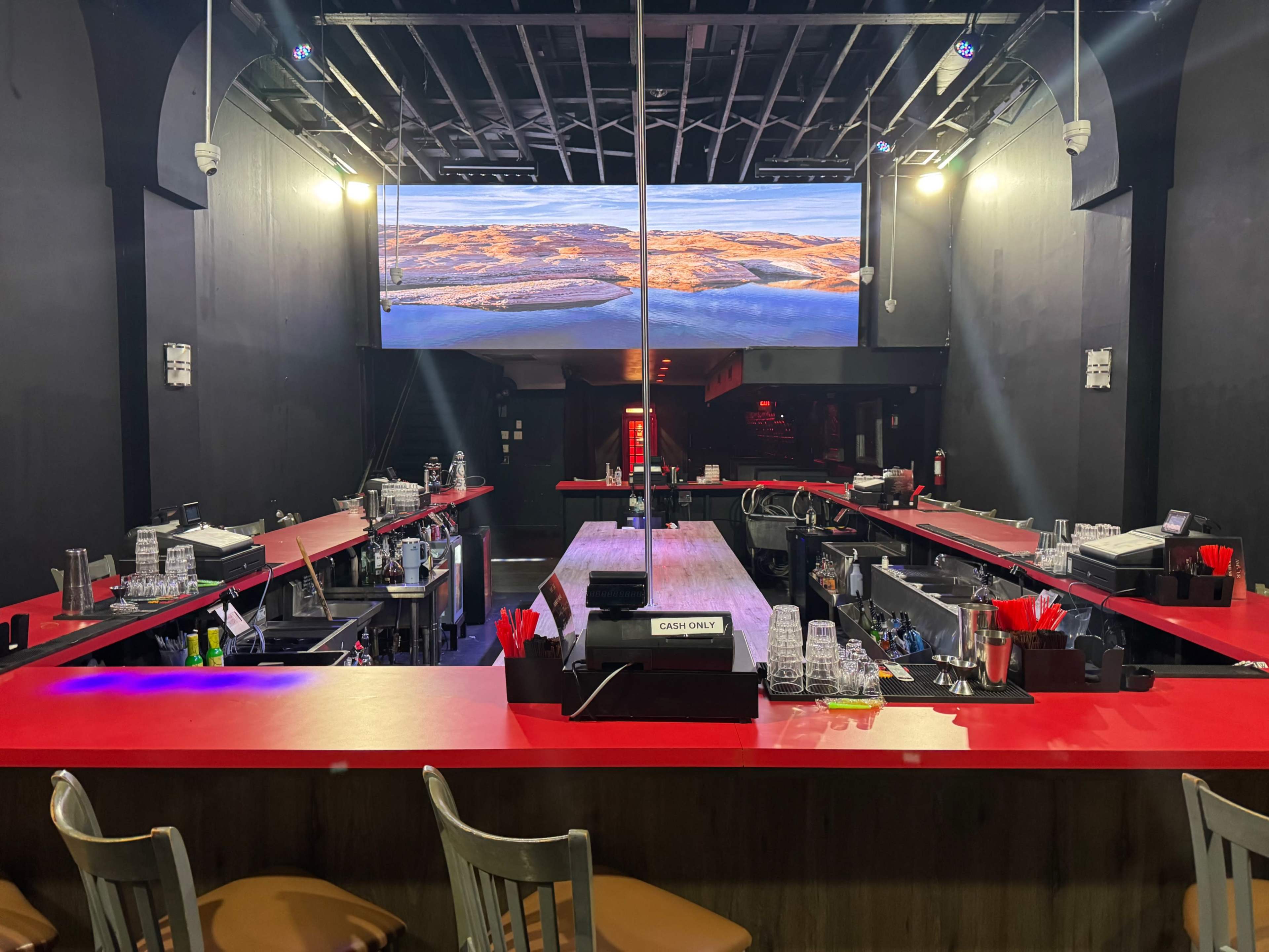 The image shows a dimly lit bar area with long red countertops, a central stage with a pole, and a large screen displaying a landscape in the background.
