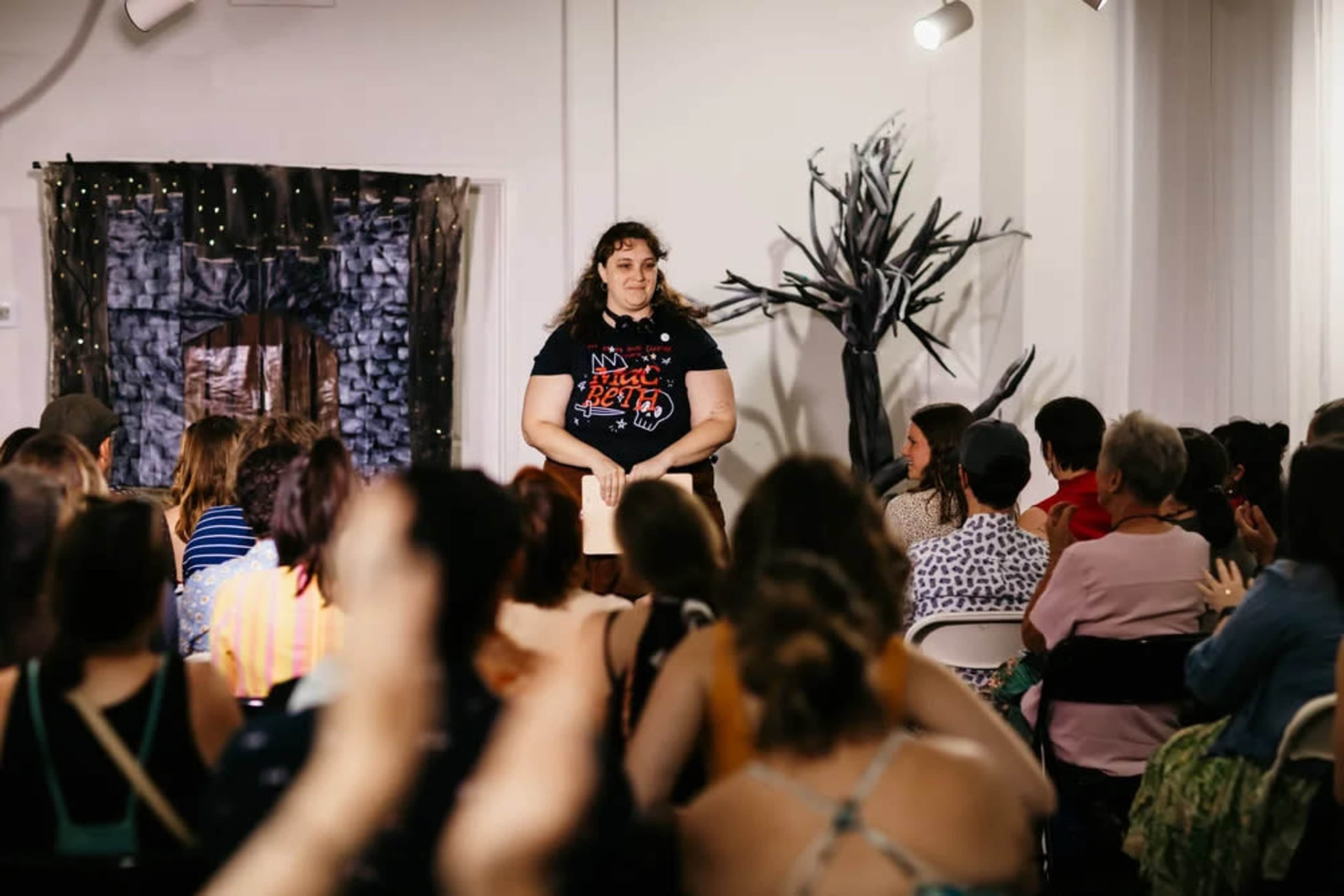 A speaker stands on stage in front of an audience, with decorations of trees and a backdrop that resembles a house.