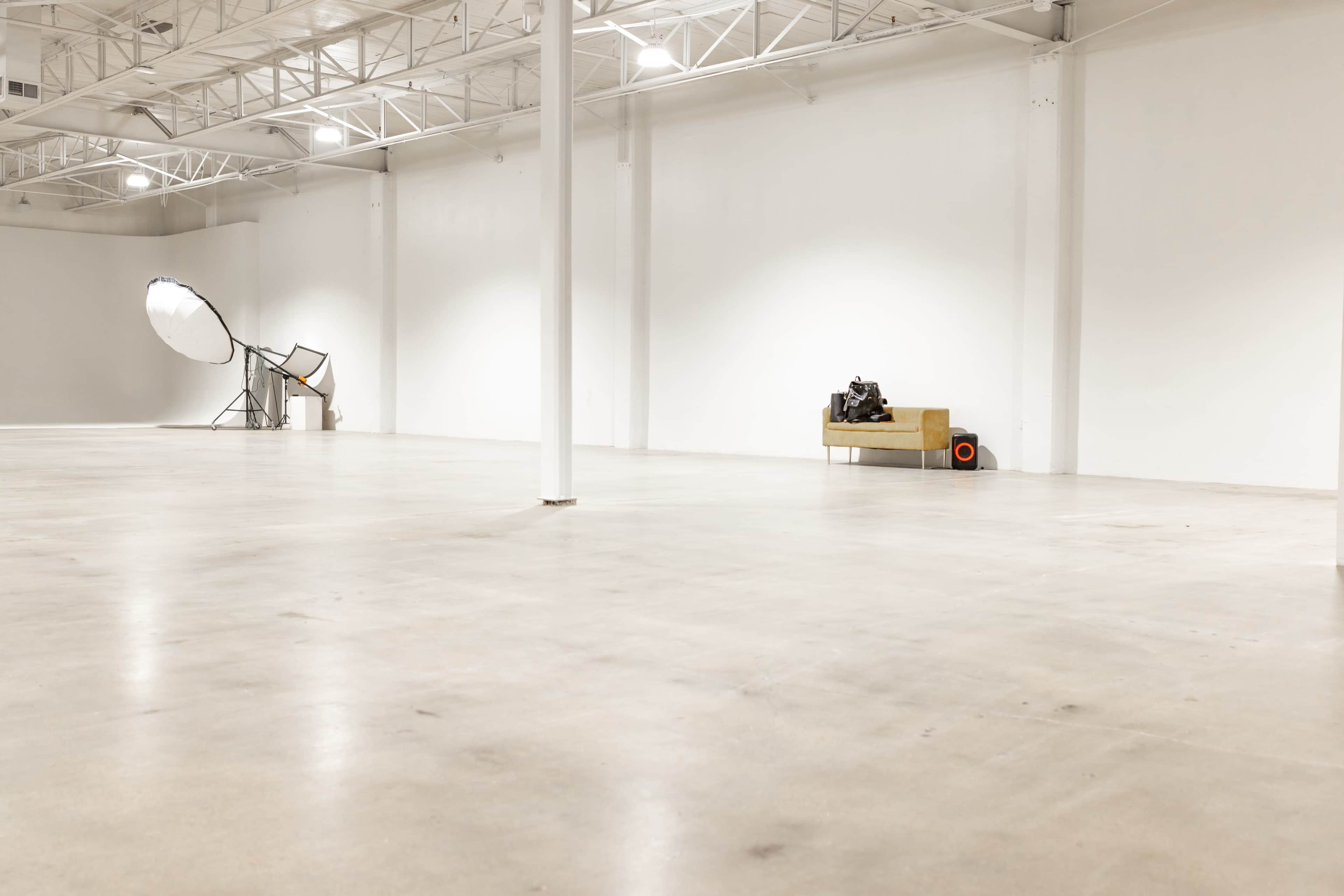 An empty industrial space with a polished concrete floor, a sofa against one wall, and large lighting equipment in the background.