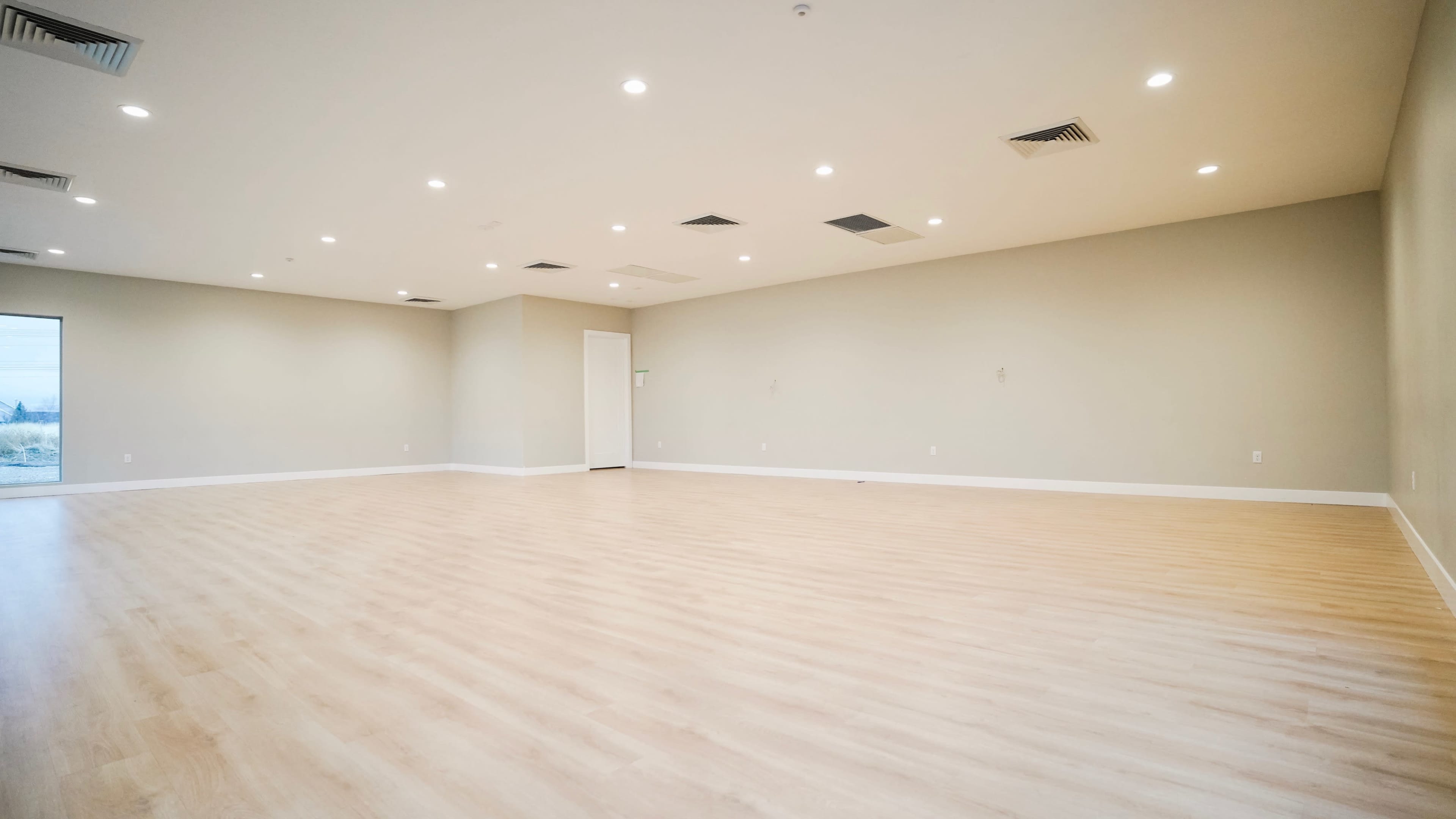 A large, empty room with light wooden flooring and neutral-colored walls, featuring recessed lighting and one door.