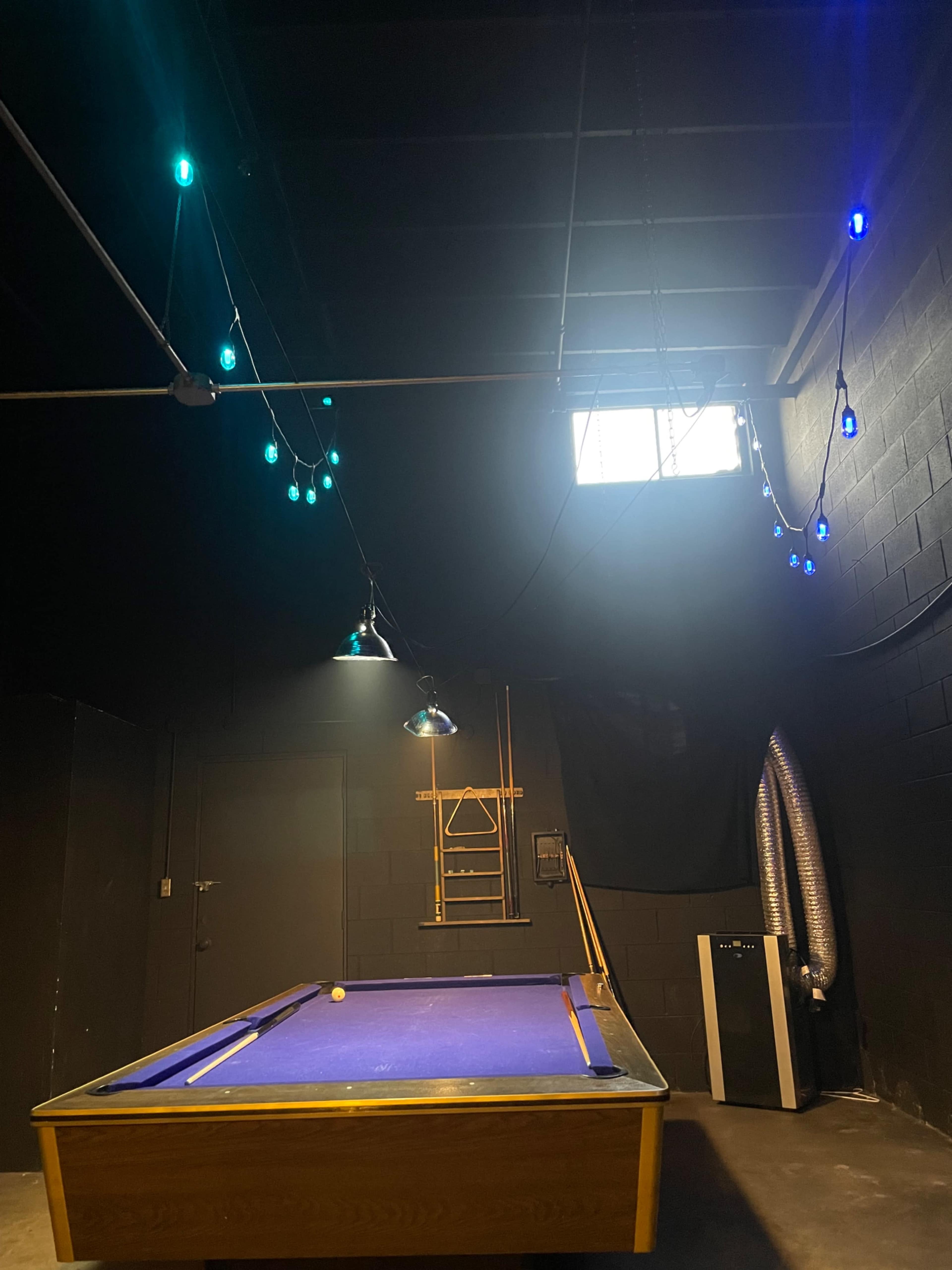 The image shows a pool table in a dimly lit room with colorful string lights overhead and a window on the wall.