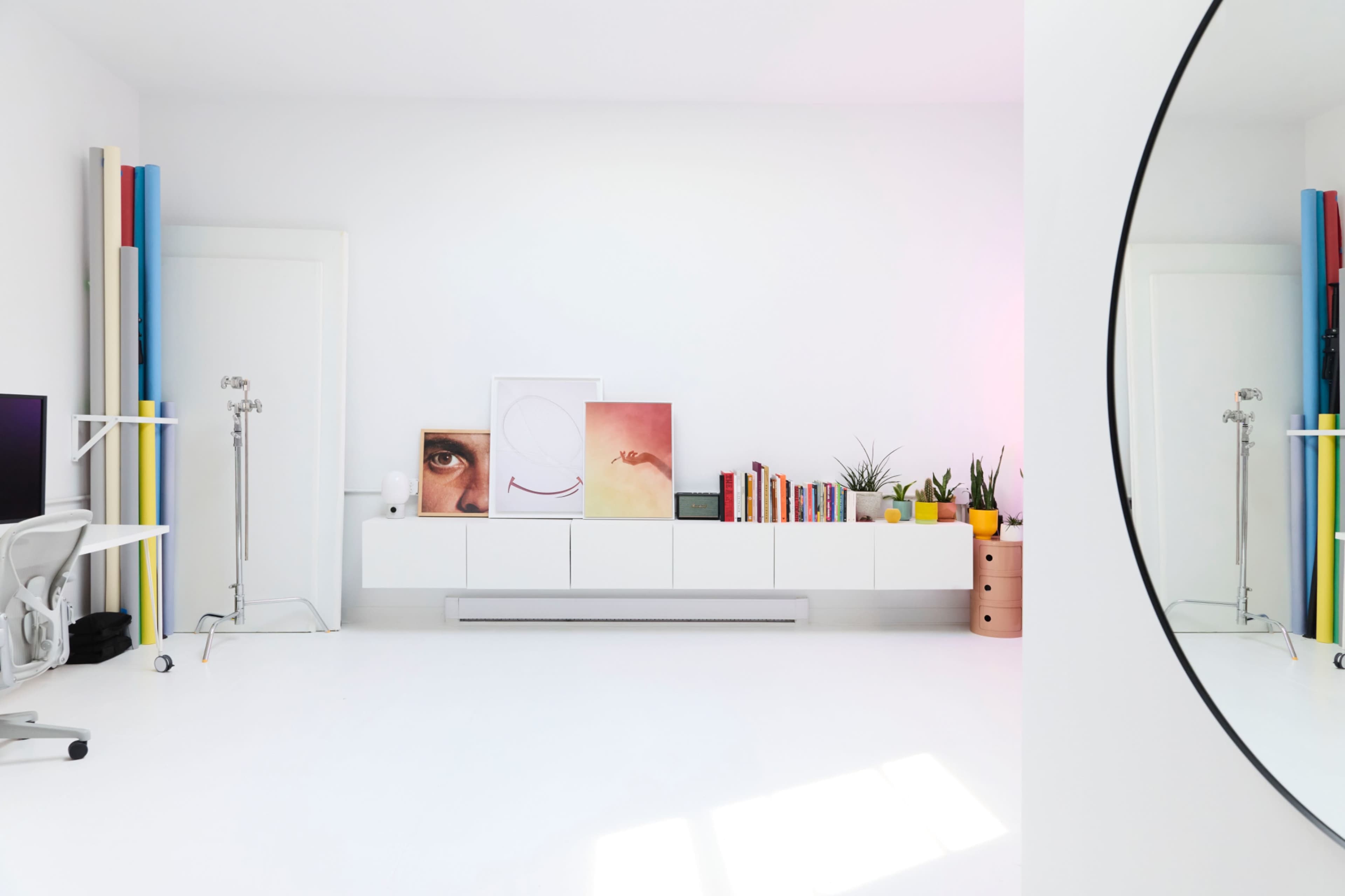 The image shows a minimalist room with a white floor and walls, featuring a shelf displaying art, books, and plants, alongside a mirror and colorful vertical panels.