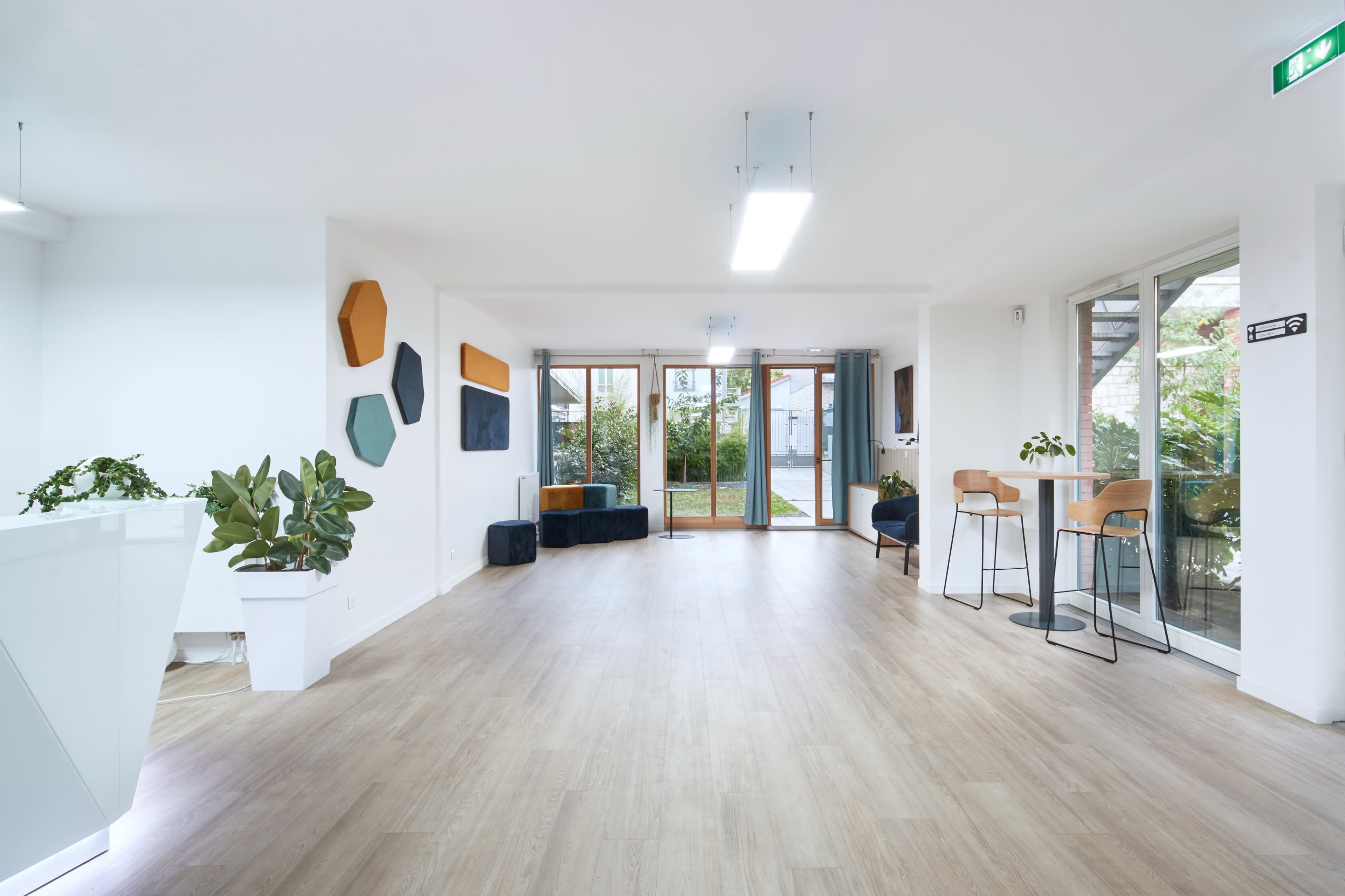 A spacious, well-lit interior features wooden flooring, modern seating arrangements, and large glass doors leading to an outdoor area.