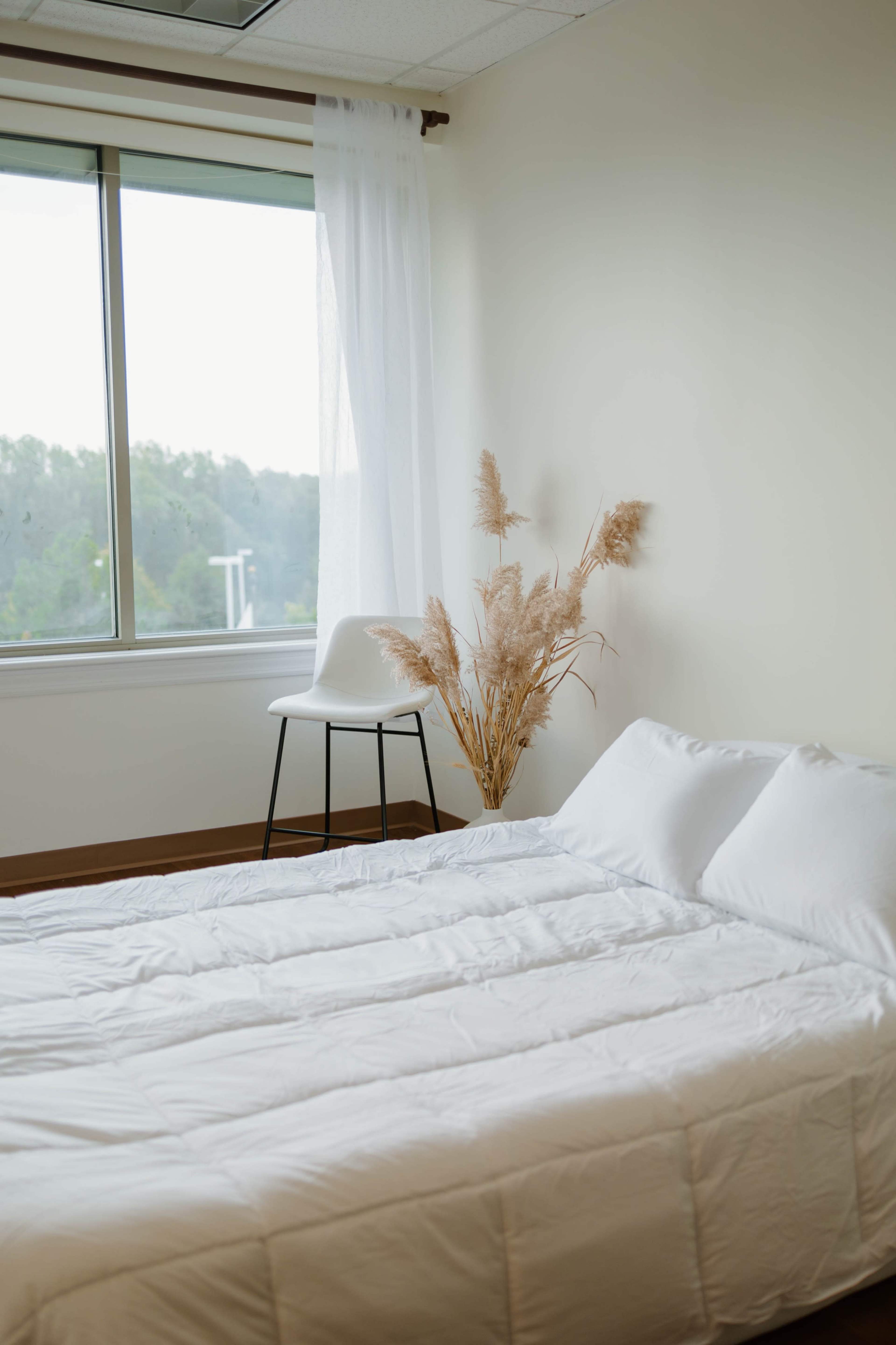 A minimalist bedroom features a bed with white bedding, a chair, and dried plants near a large window overlooking greenery.