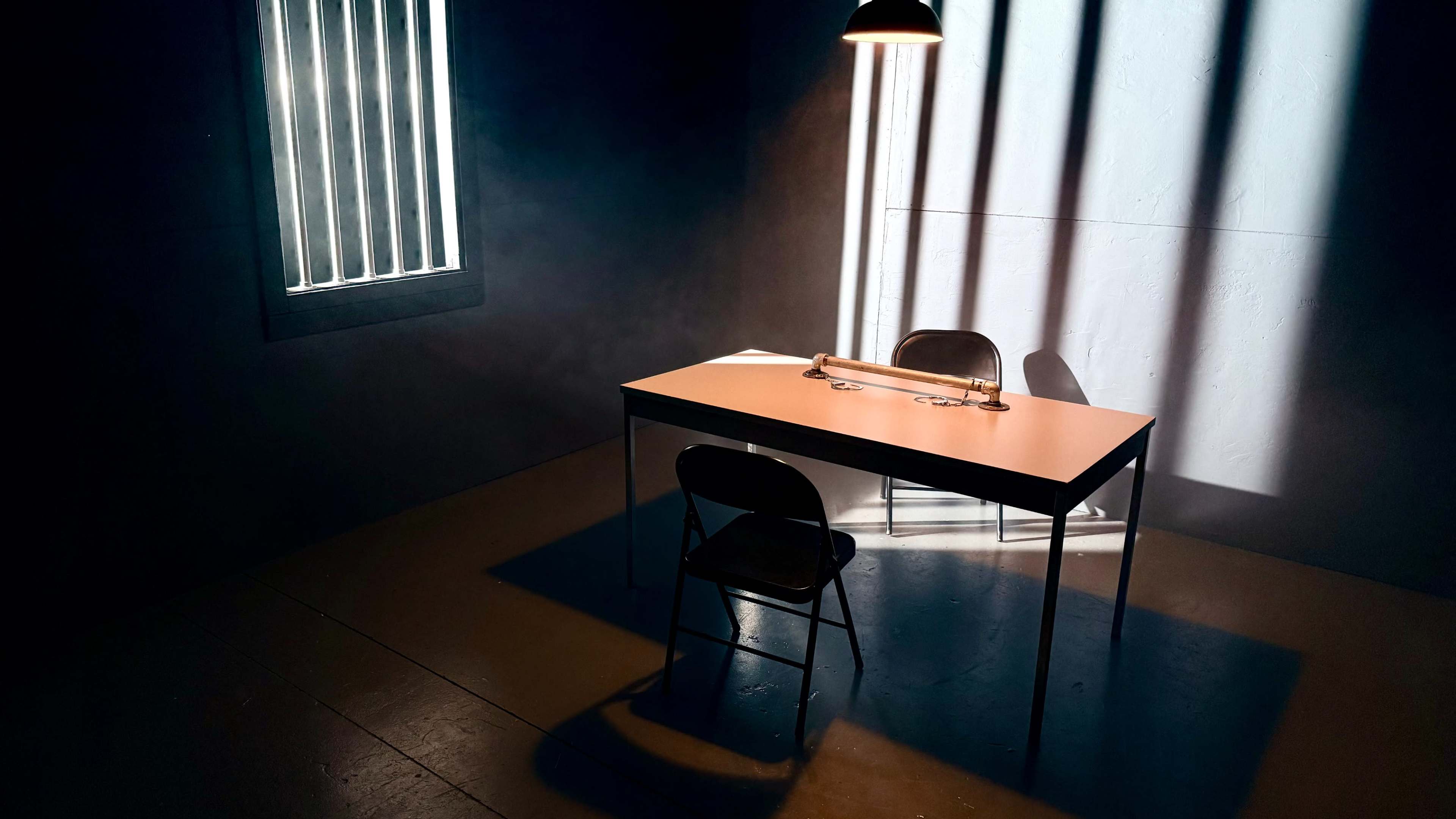 A wooden table with a metal bar sits under a single overhead light, accompanied by a black chair, in a dimly lit room with barred windows.