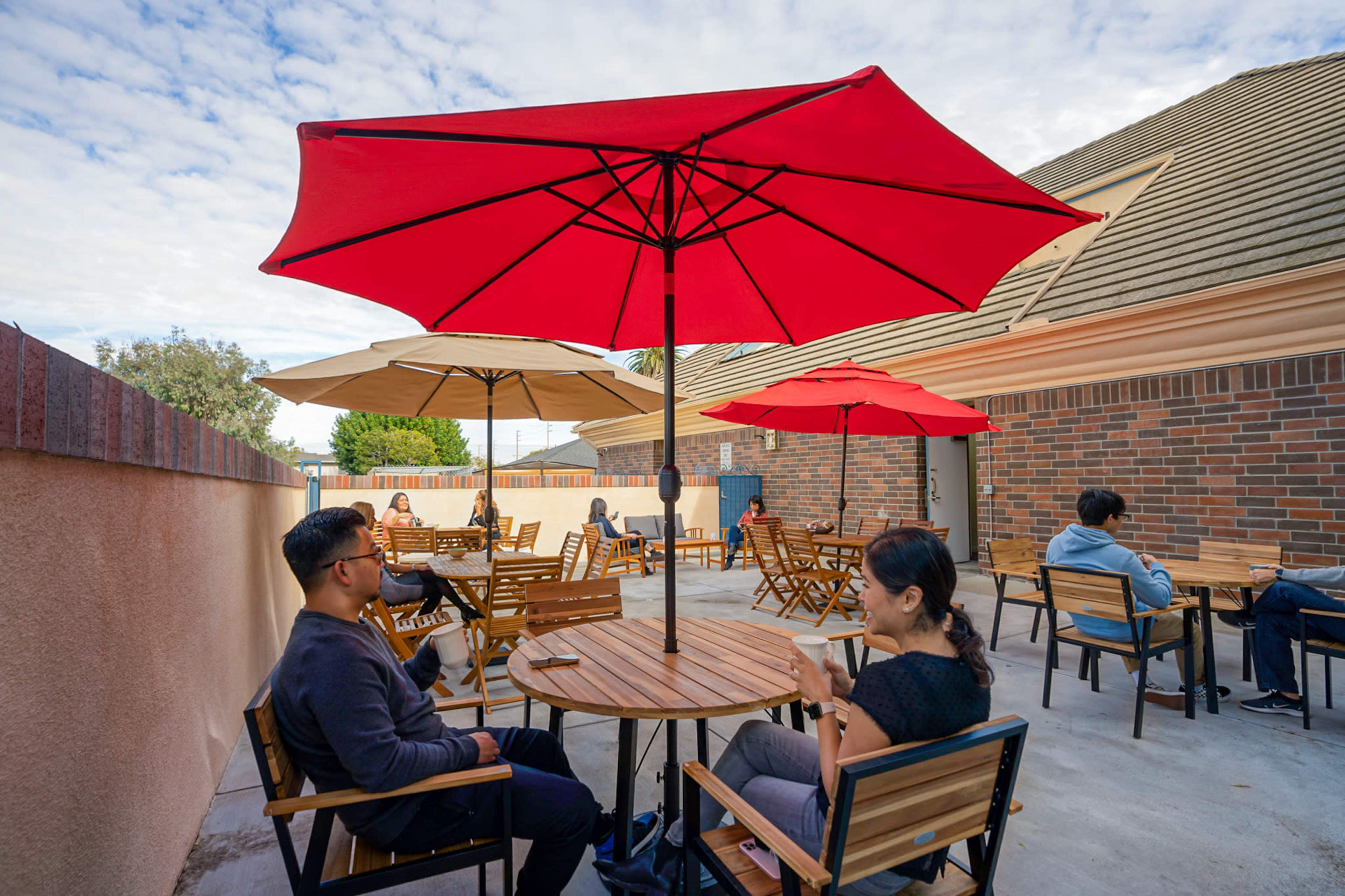 A group of people sits at wooden tables under red umbrellas in an outdoor patio area.