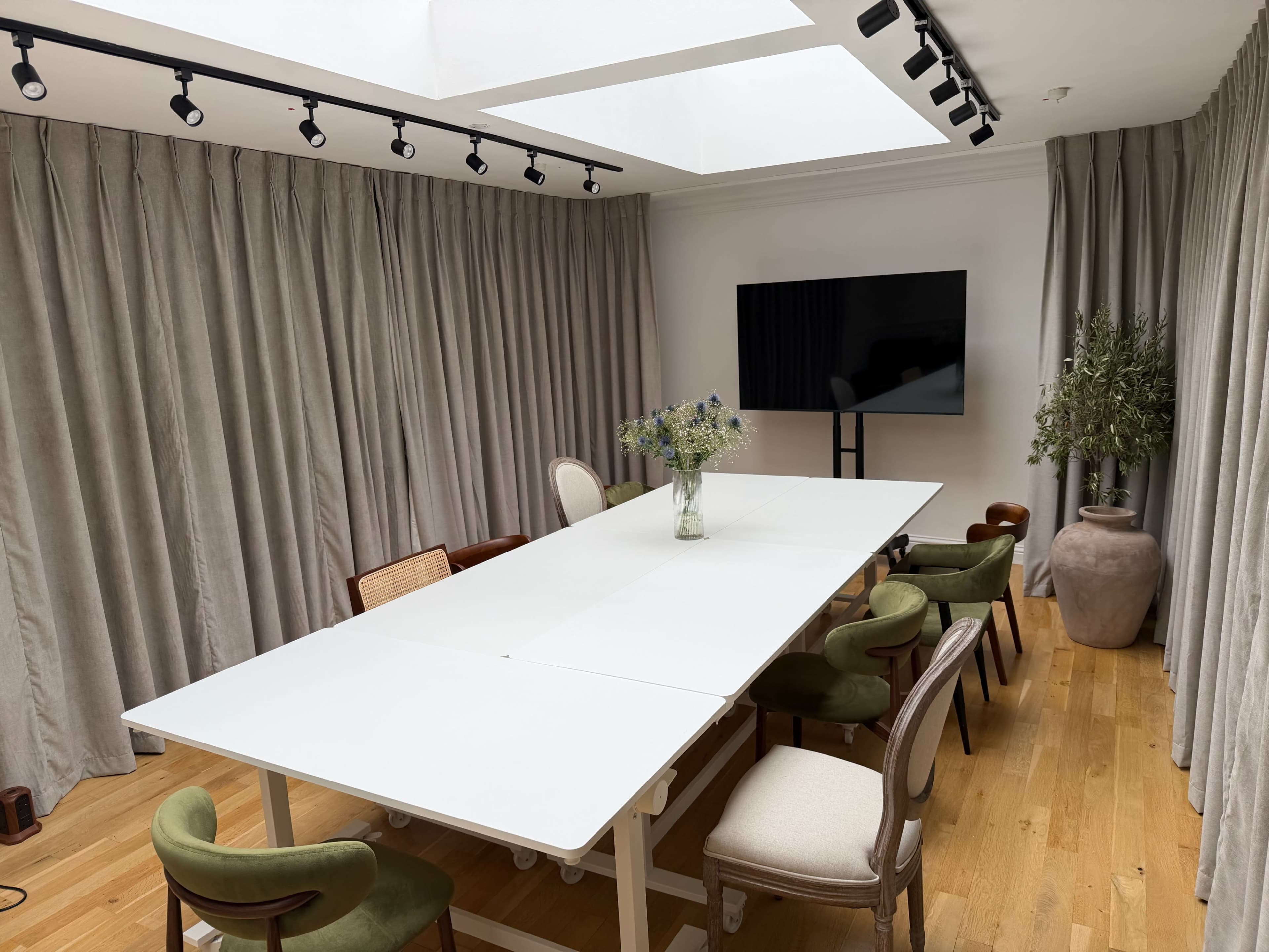 A spacious meeting room features a long white table surrounded by several chairs, with large curtains, a television, and a decorative plant.