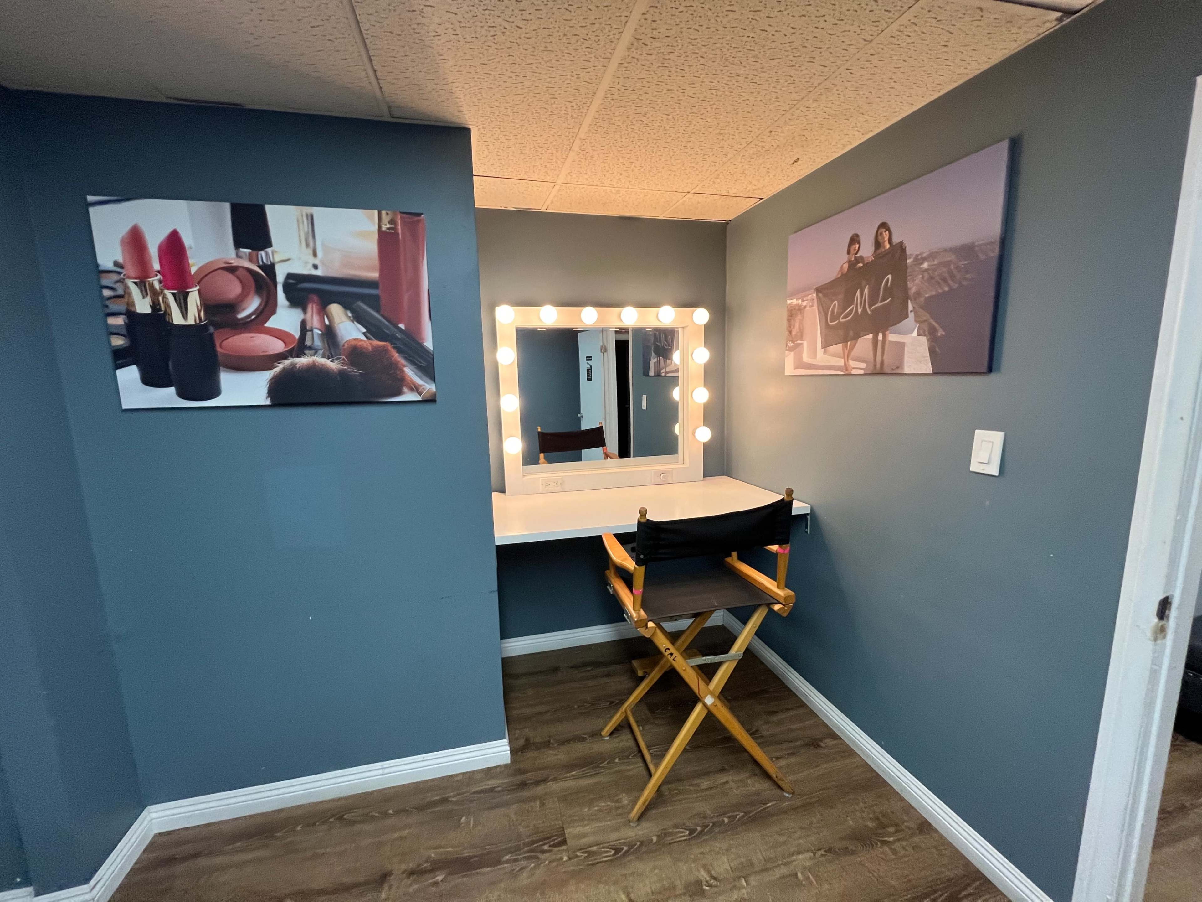 A small room with a makeup vanity featuring a lighted mirror, a director's chair, and wall art depicting cosmetics and a group of people.