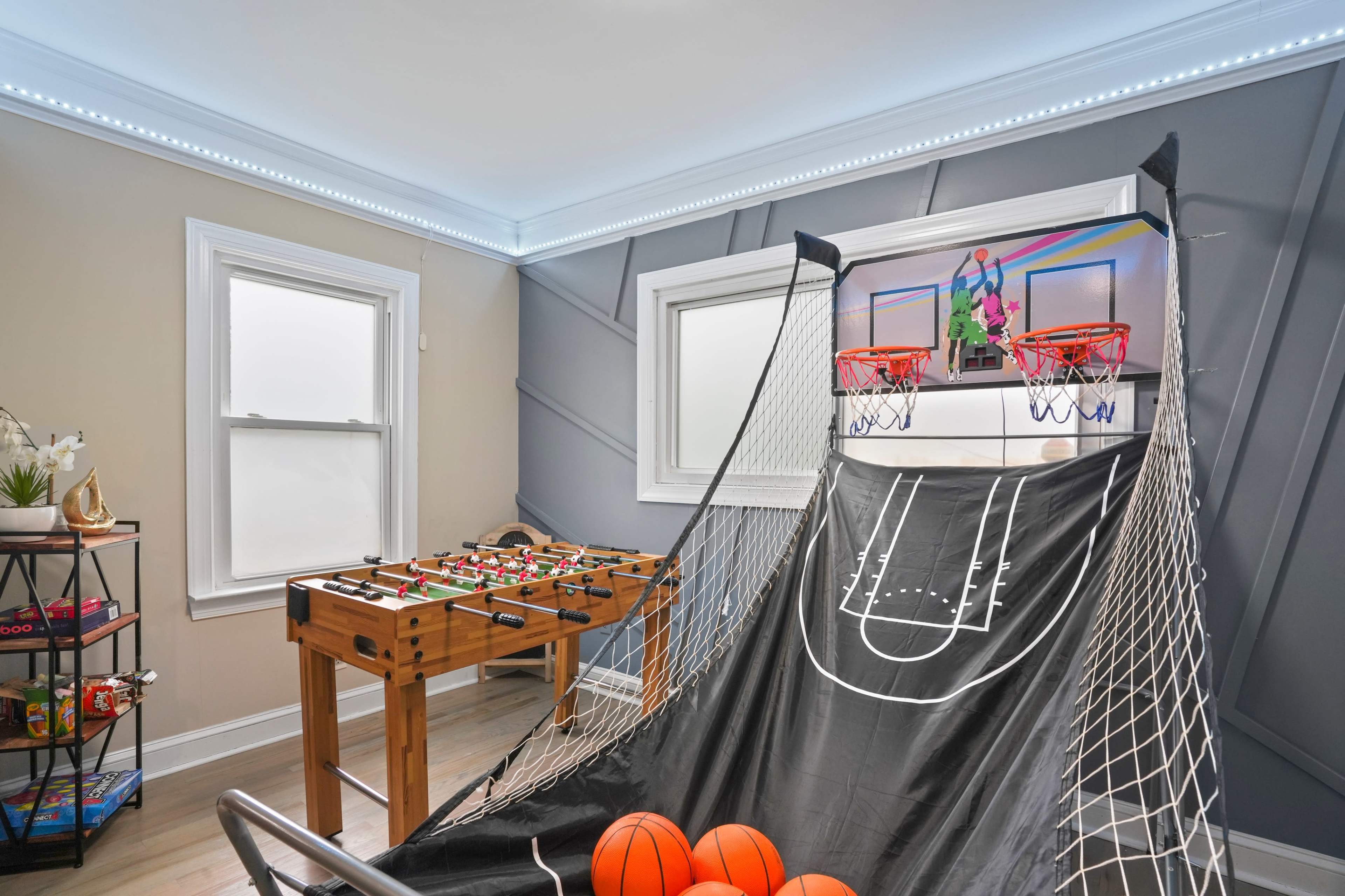 A game room featuring a basketball shooting game and a foosball table beside it, with bright lighting along the ceiling and windows allowing natural light to enter.