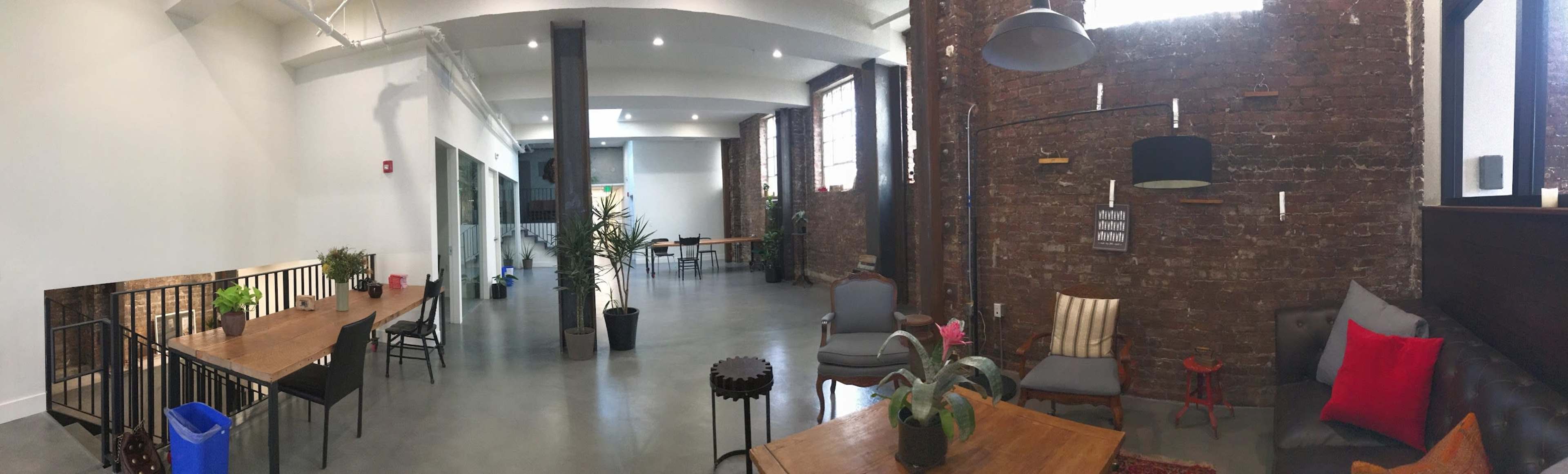The image shows a spacious, modern interior with exposed brick walls, wooden furniture, and plants arranged in a casual office or communal space.
