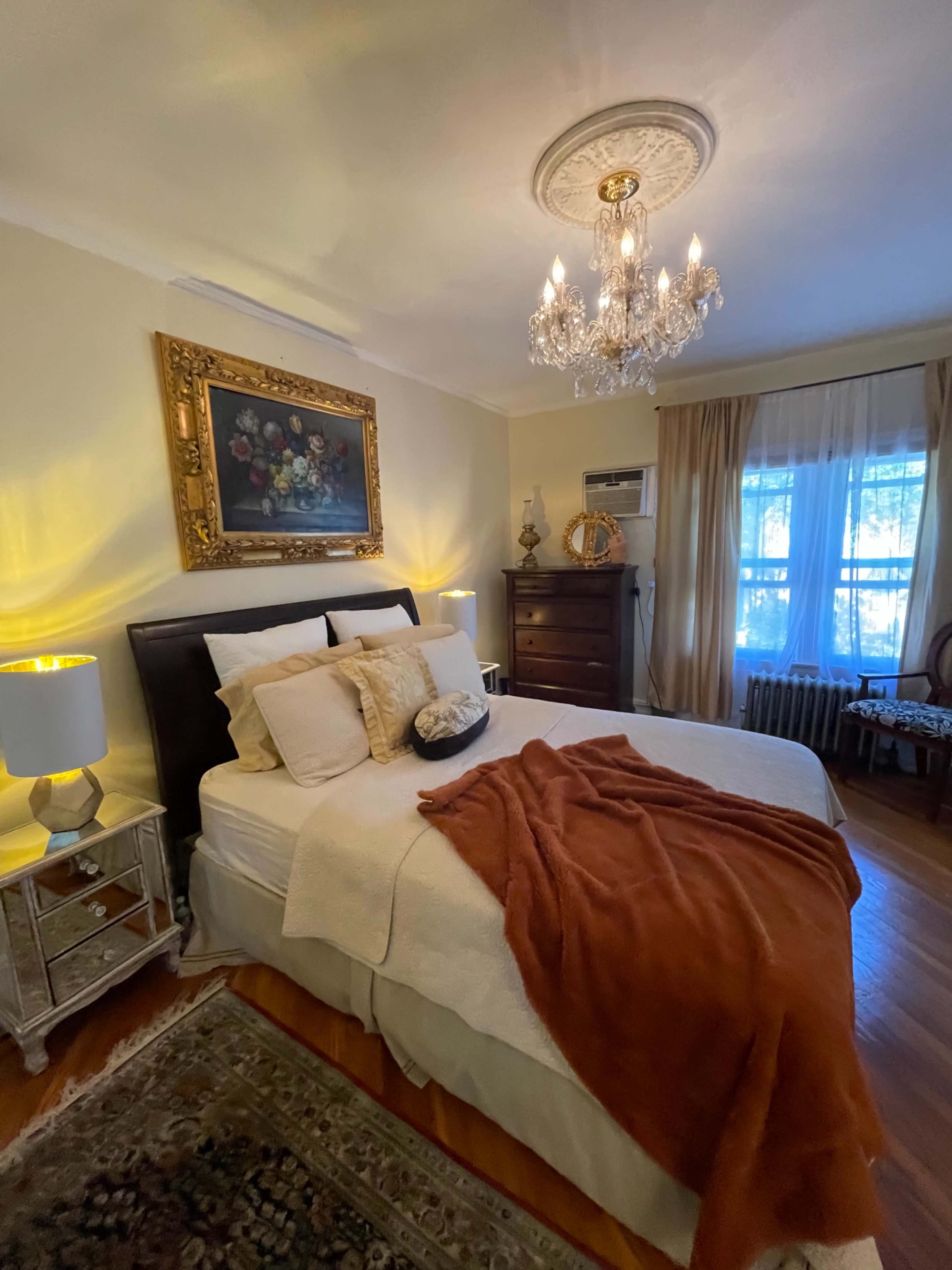 A cozy bedroom featuring a bed with white linens, a warm throw, a chandelier, and a window with sheer curtains.