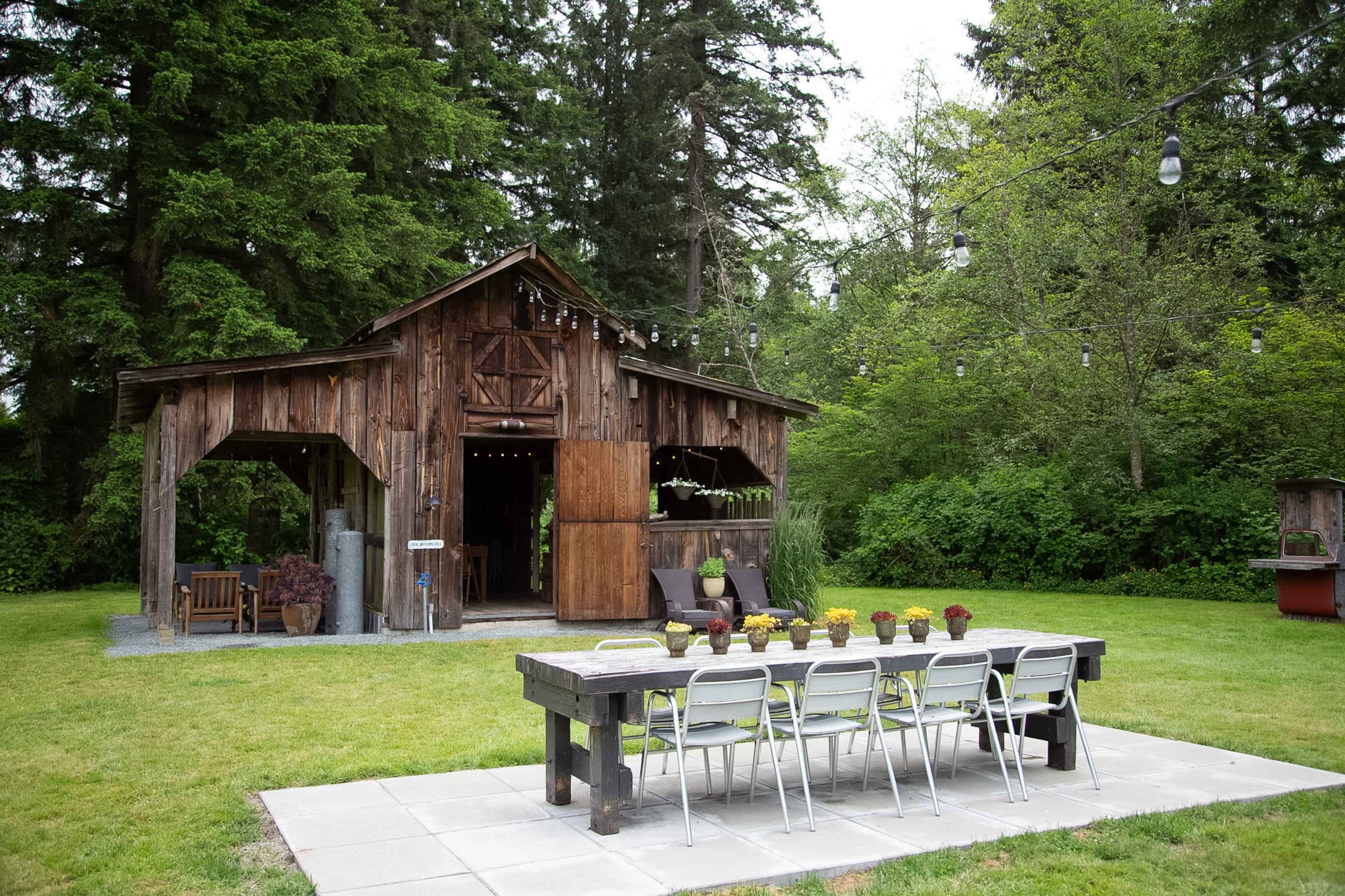 A rustic wooden barn stands in a green yard, accompanied by a long dining table set with potted plants and surrounded by chairs.