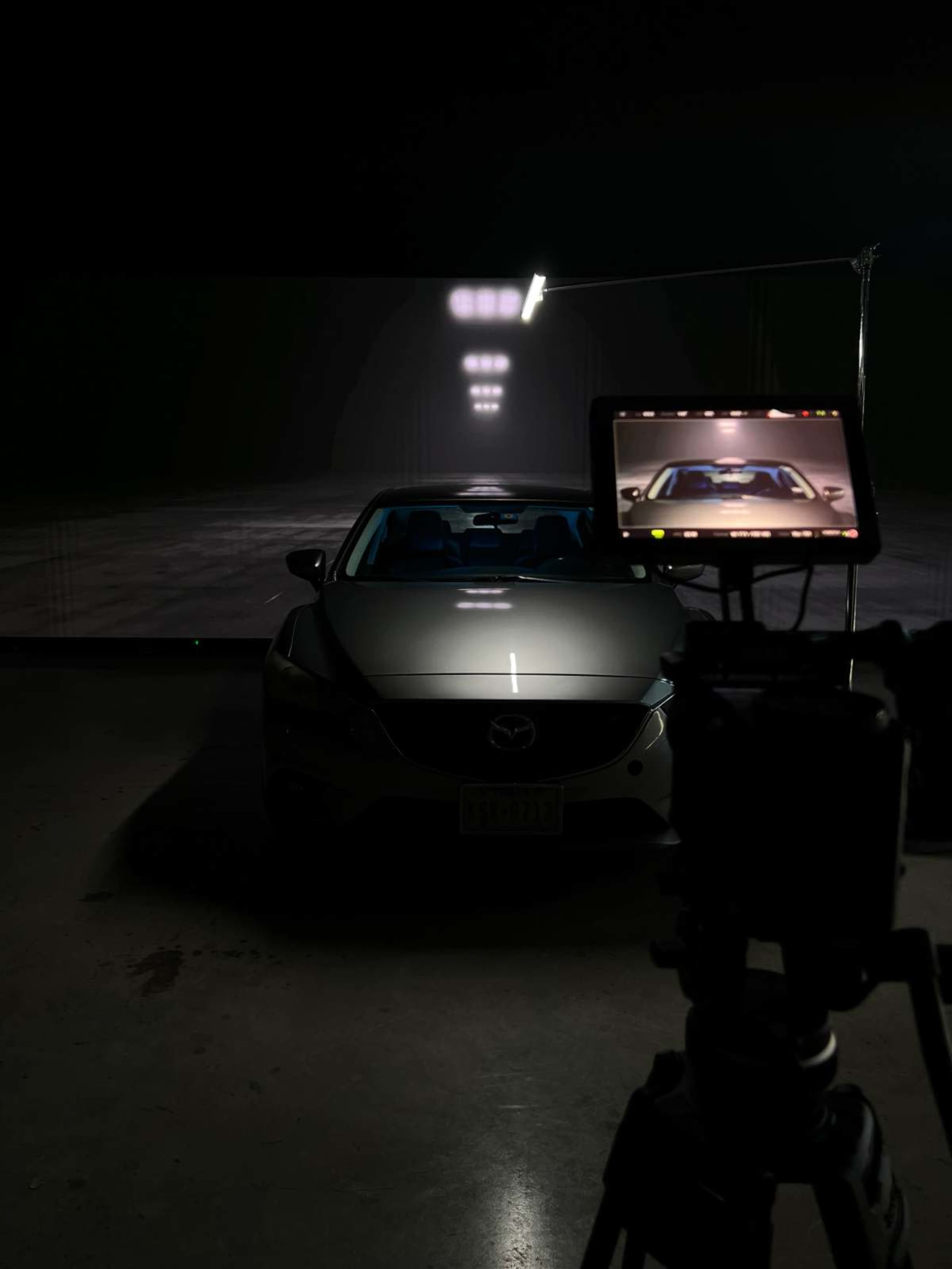 A silver car is positioned in front of a backdrop with bright lights, while a camera is focused on it from a nearby stand.