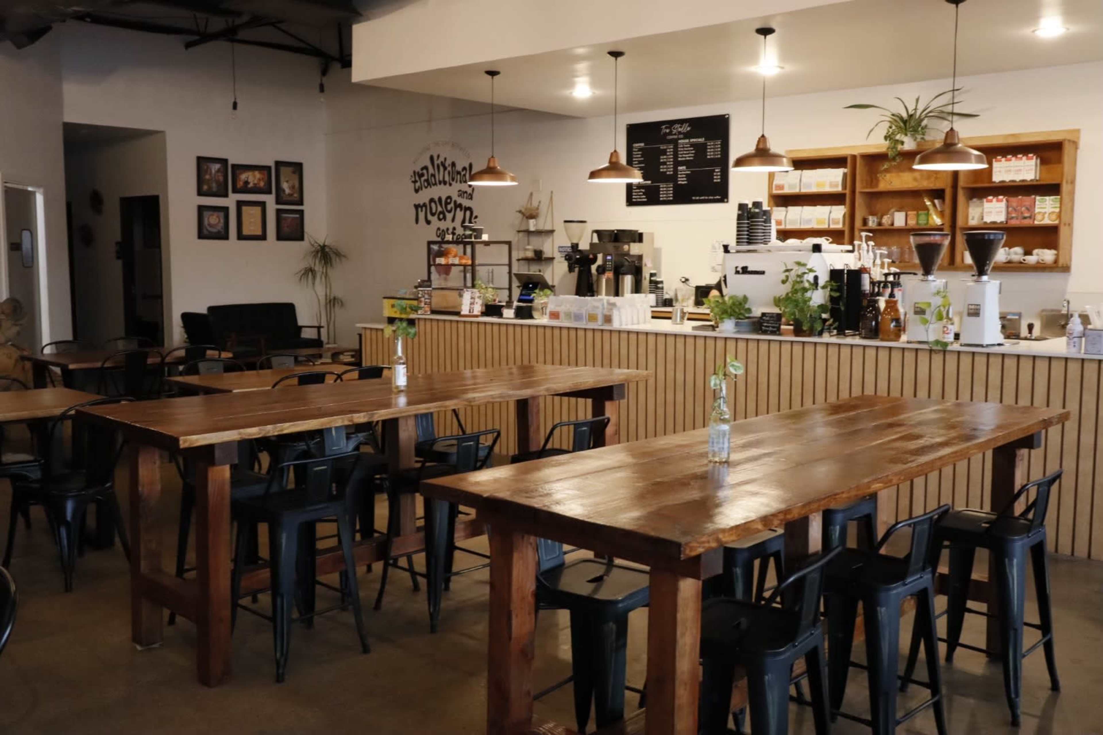 The image shows a modern coffee shop interior with wooden tables, black chairs, and a service counter featuring various coffee-making equipment and products.