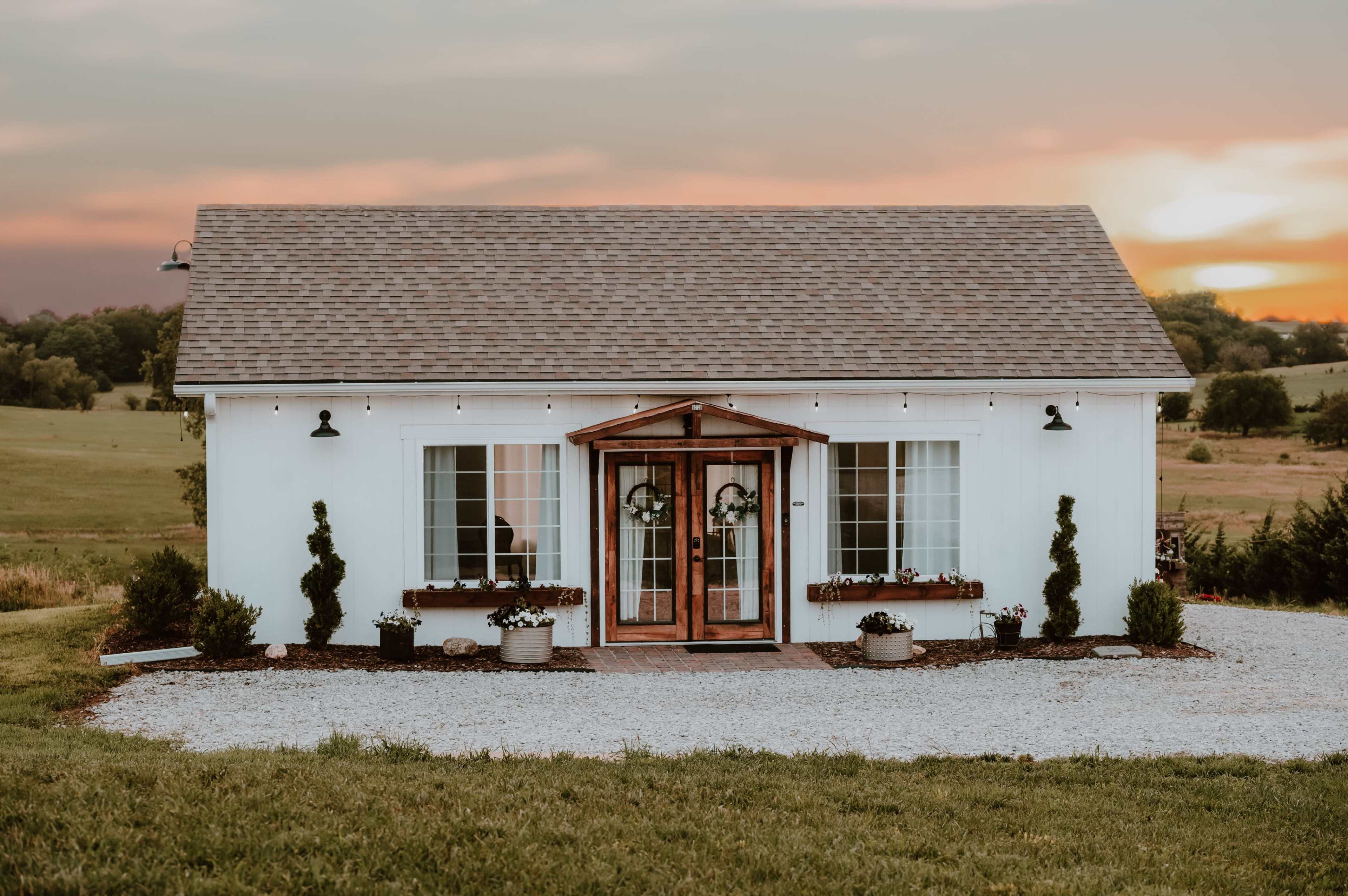 A small, white cottage with a wooden porch and two symmetrical entrance doors is set against a sunset backdrop.