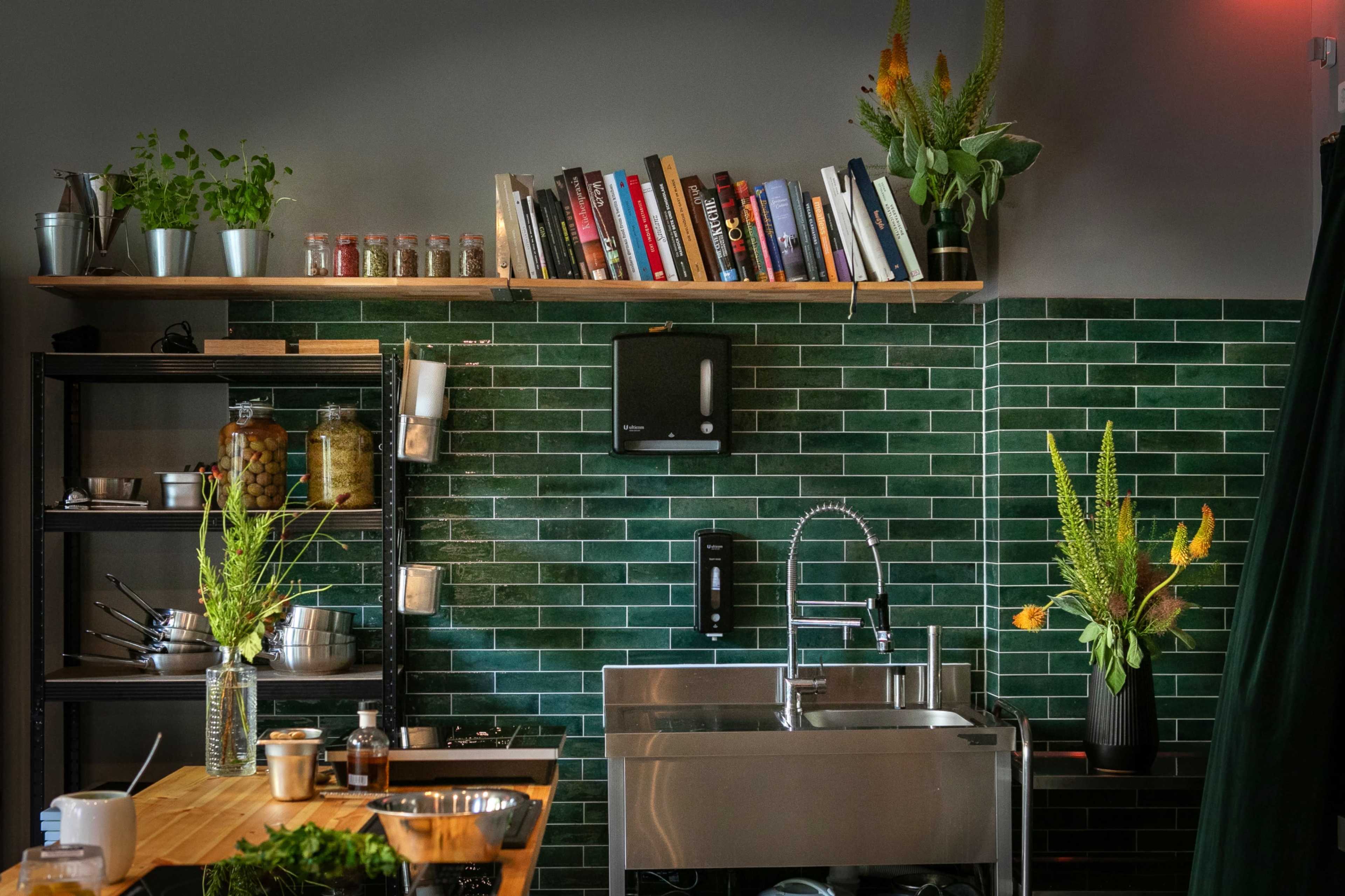 The image shows a modern kitchen with green tiled walls, a stainless steel sink, and open shelving displaying books and plants.