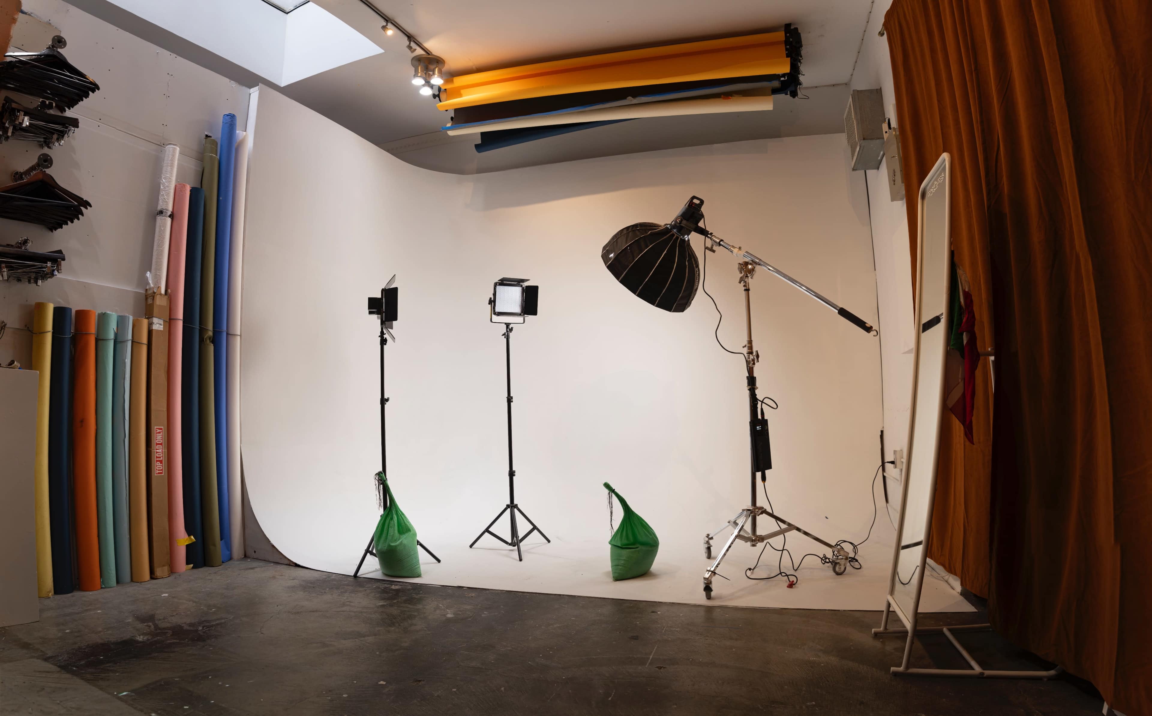 The image shows a photography studio setup featuring a seamless backdrop, two studio lights on stands, and a large softbox, along with colorful rolls of backdrops visible on one side.
