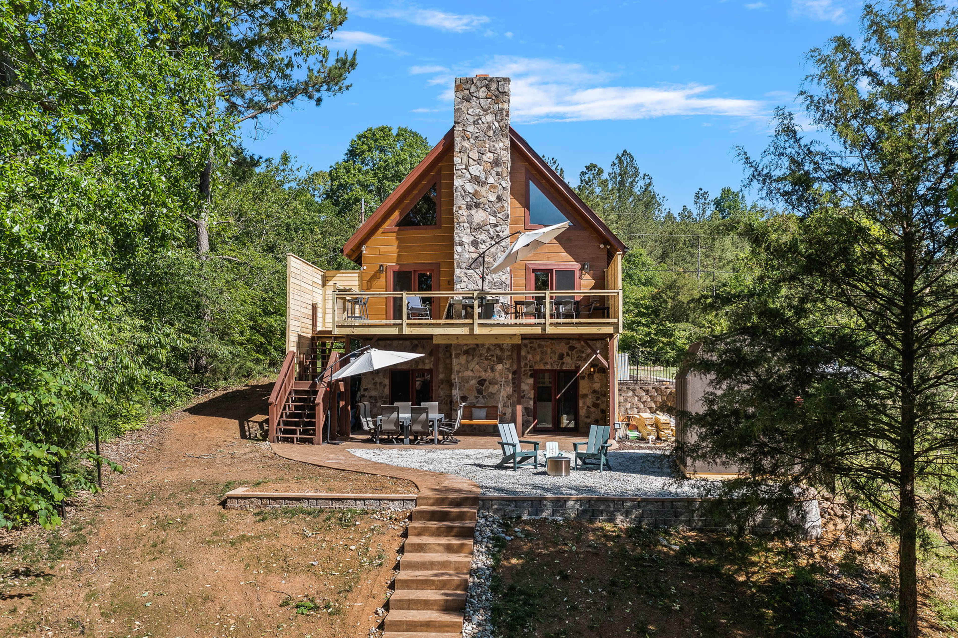 A three-story house with a stone chimney and a wooden deck is situated among trees, featuring outdoor seating and a gravel pathway.