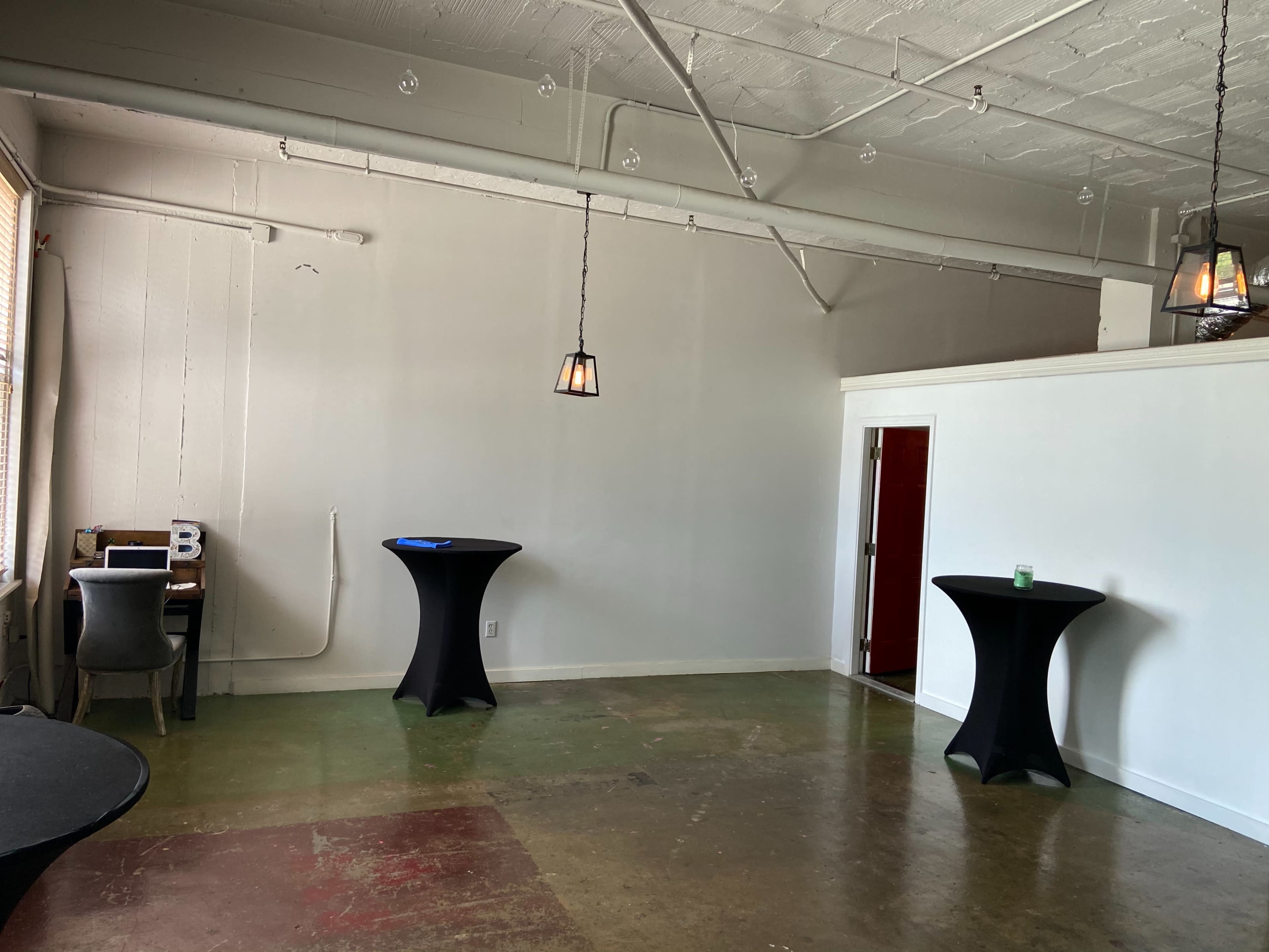 A spacious room with a polished concrete floor, two black cocktail tables, a small desk with equipment, and a doorway leading to another area.