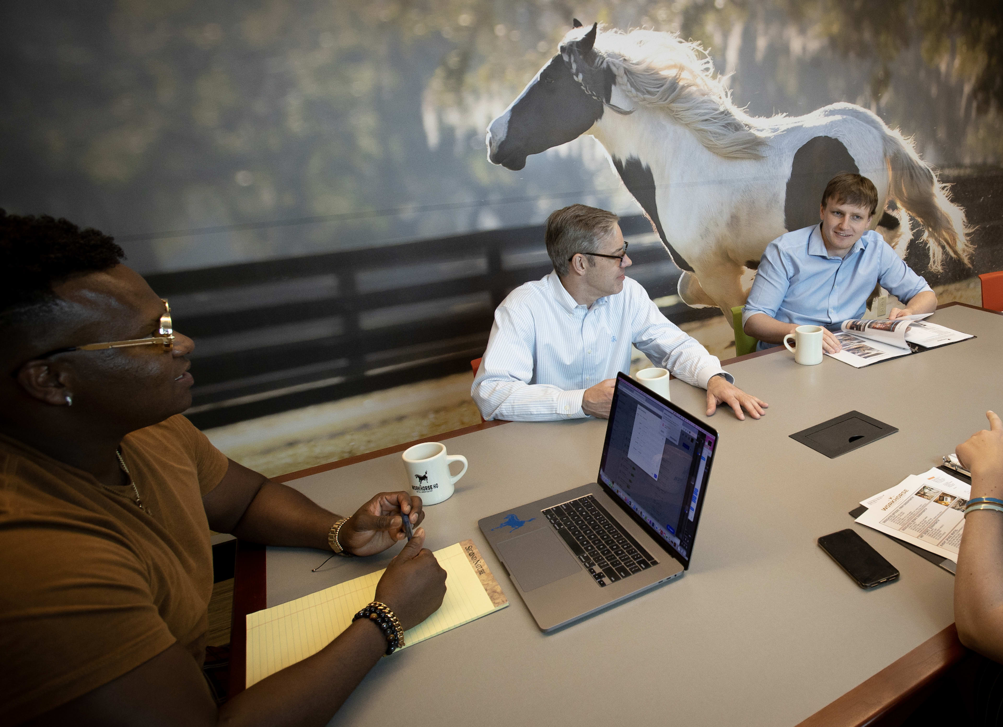 A group of three people sits around a conference table with a laptop and notebooks, while a large horse mural decorates the wall behind them.