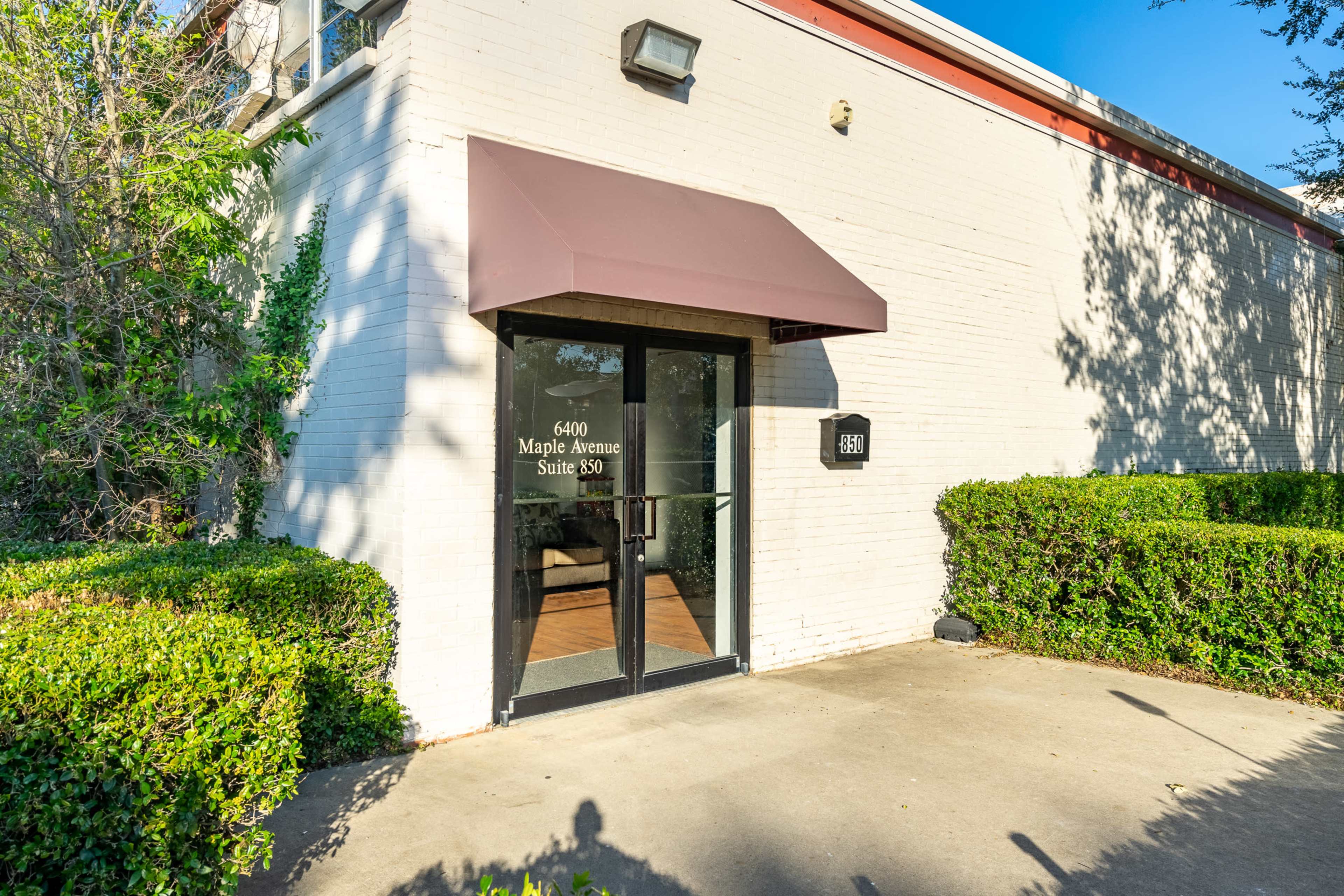 The image shows the entrance of a building featuring a glass door with a maroon awning and surrounding green shrubs.