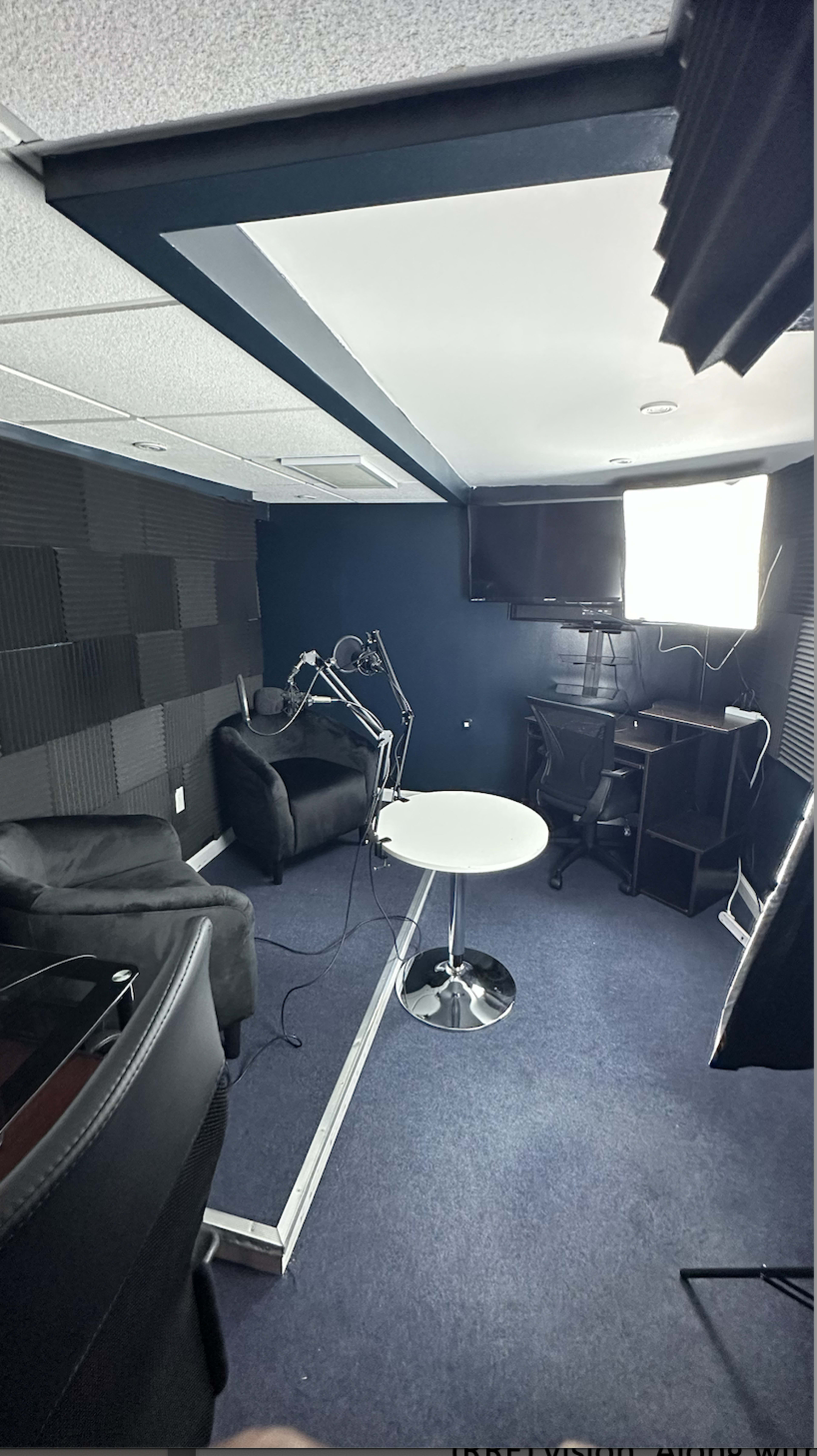 A small recording studio with black soundproofing panels, a round white table, two black chairs, and equipment including a monitor and a light.