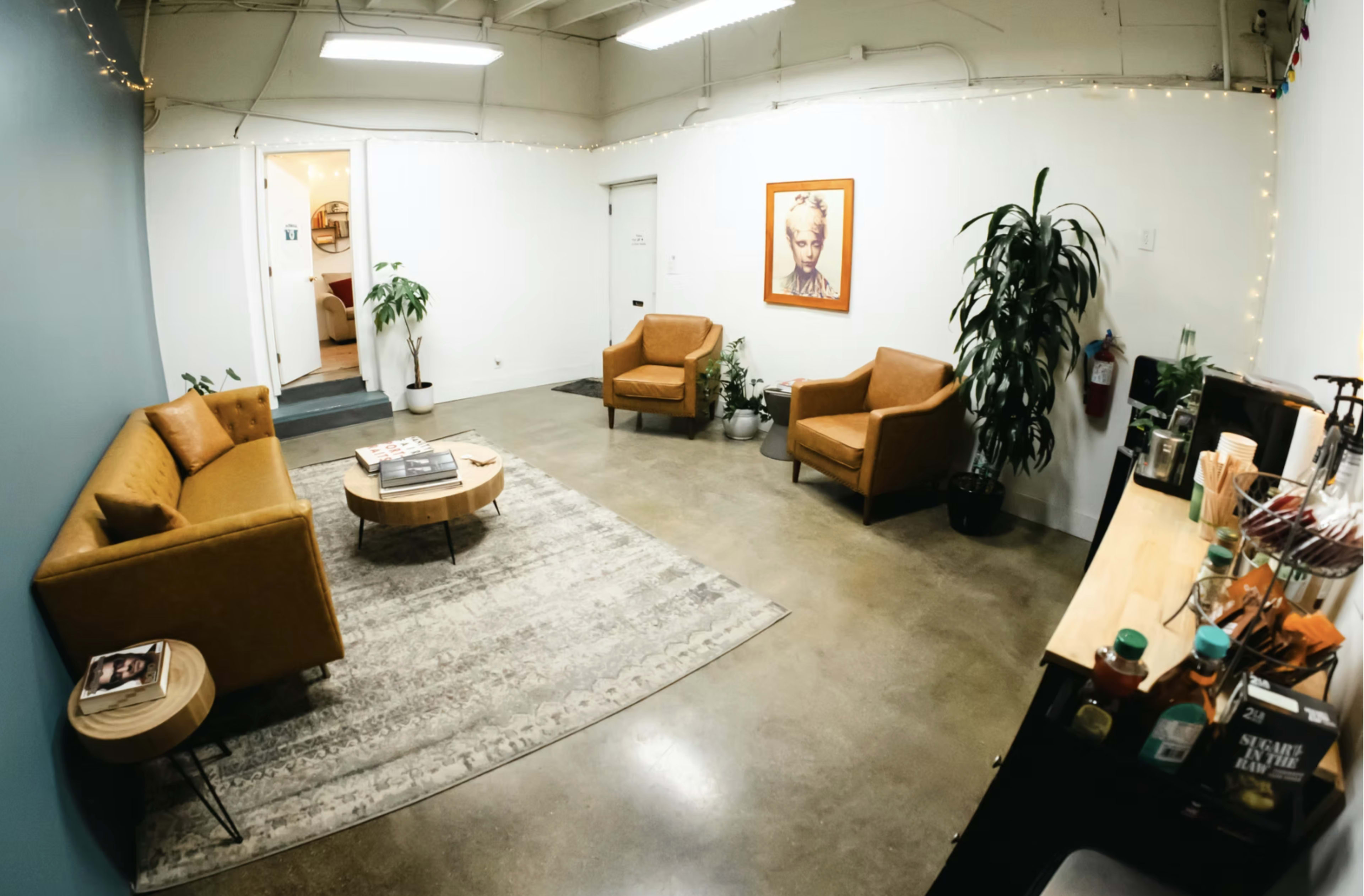The image shows a cozy lounge area with two brown chairs, a small table, and a rug, surrounded by potted plants and art on the walls.