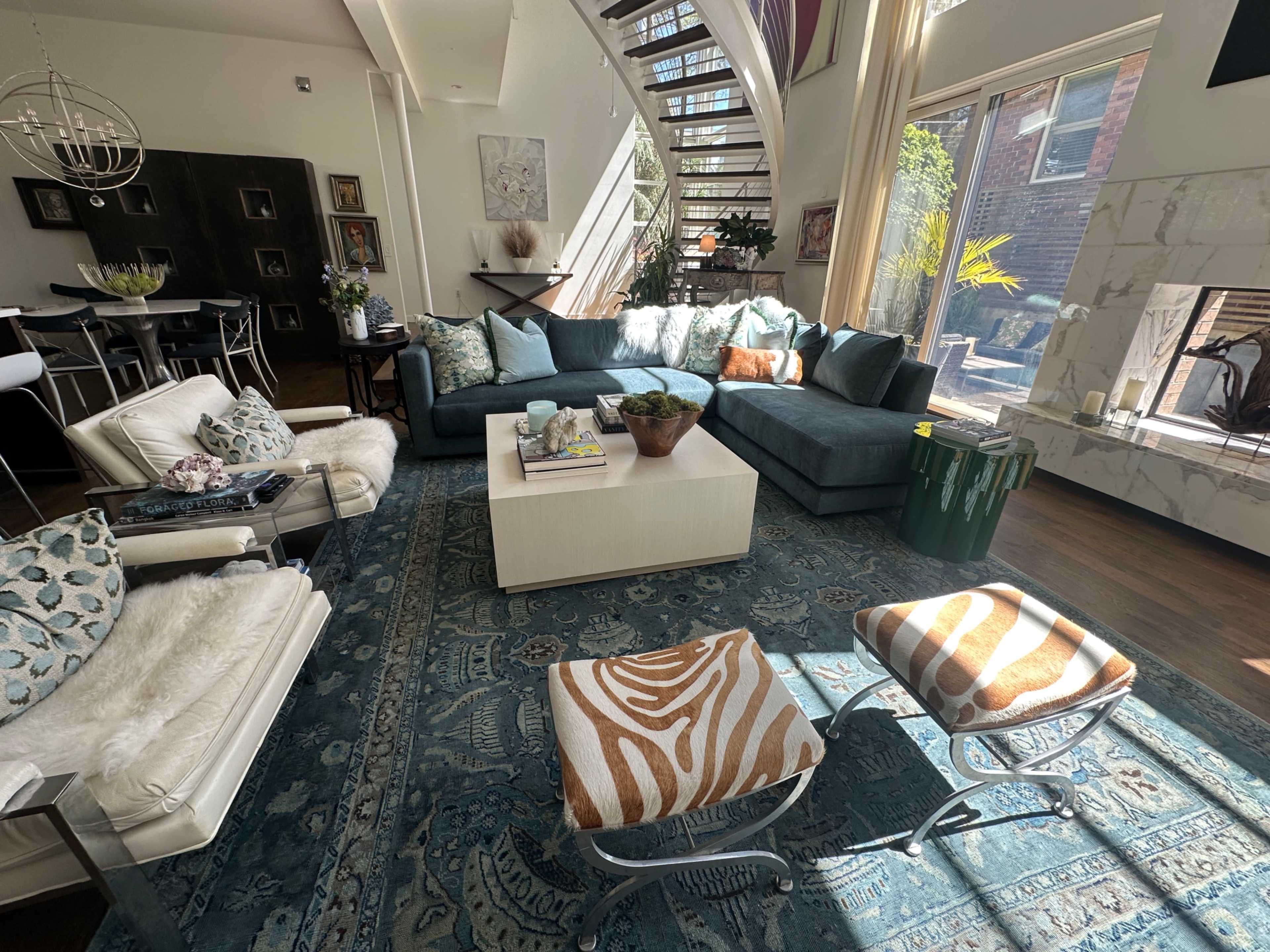 The image shows a modern living room featuring a large sectional sofa, a coffee table, and decorative chairs, with a staircase and large windows illuminating the space.