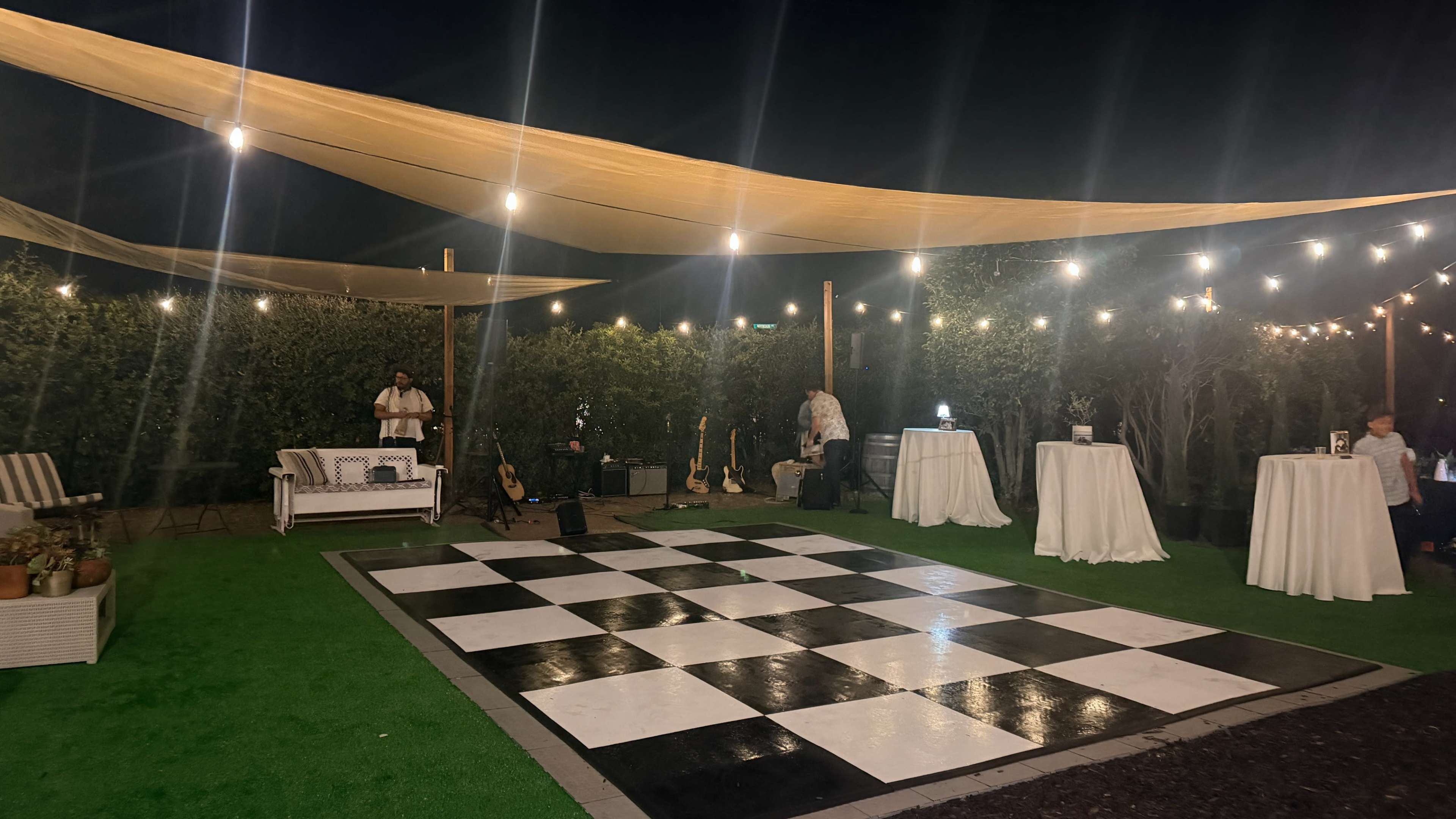 The image shows an outdoor event space featuring a black and white checkered dance floor, tables, and string lights overhead amidst greenery.