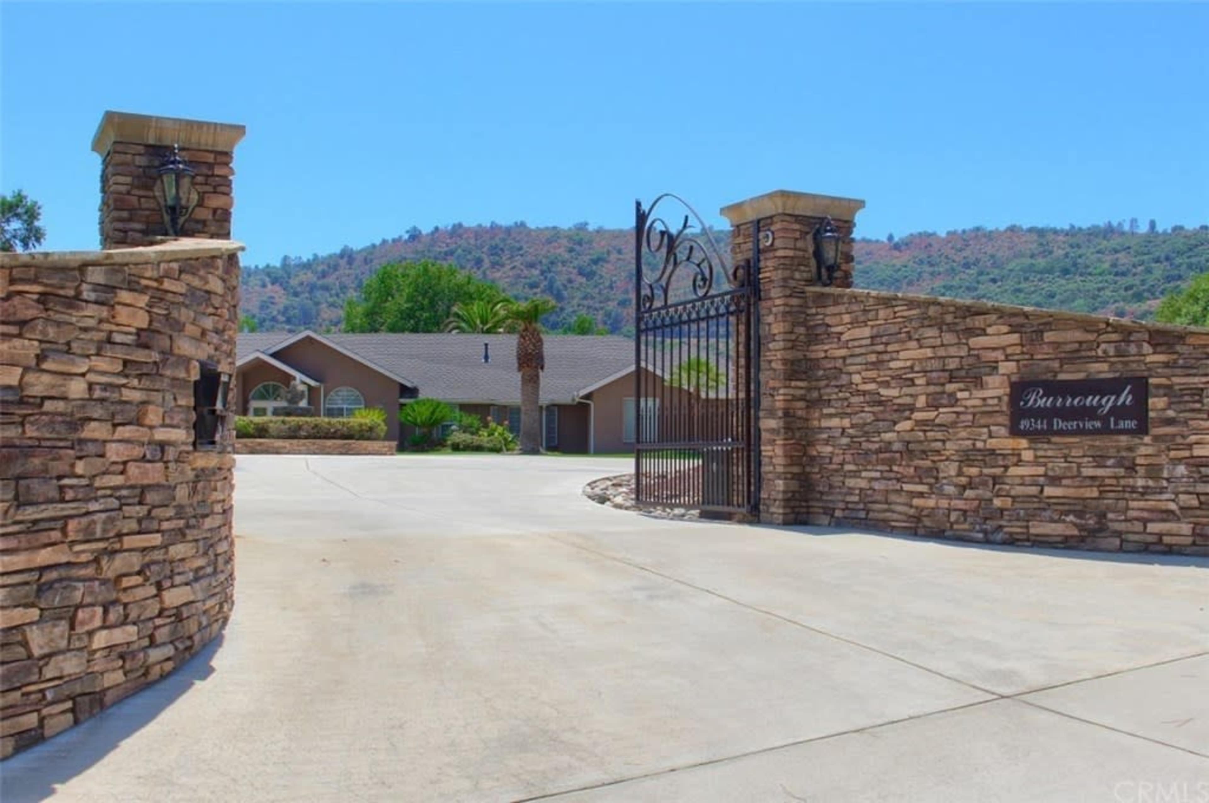 A gated entrance with stone pillars leads to a driveway surrounded by greenery and a mountainous backdrop.