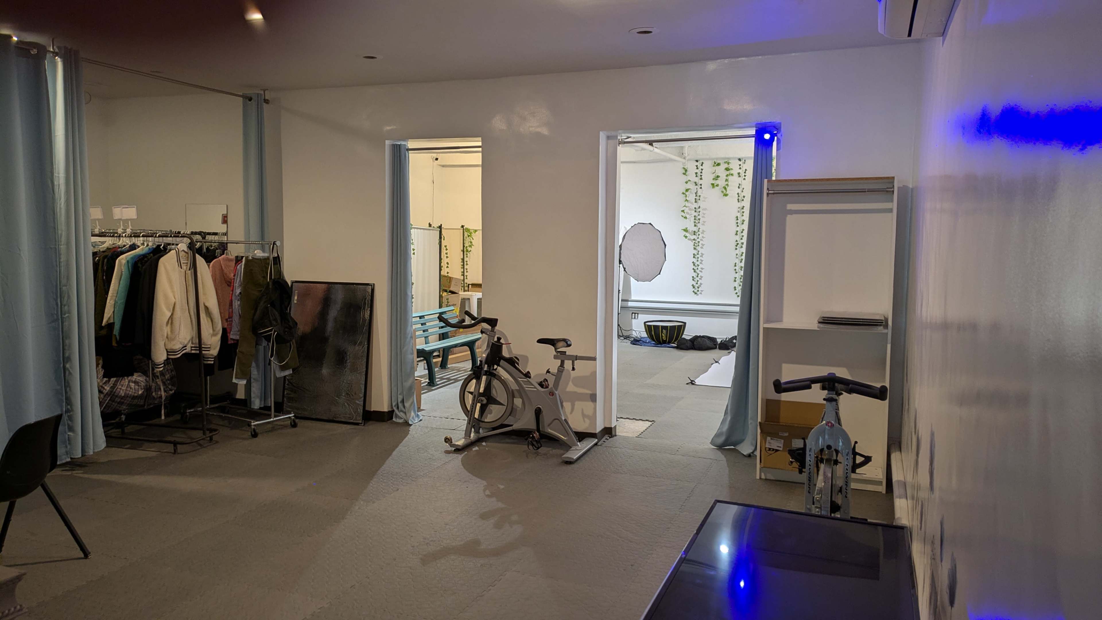 The image shows a spacious room with clothing racks on one side, a stationary bike in the center, and two doorways leading to a well-lit area in the background.