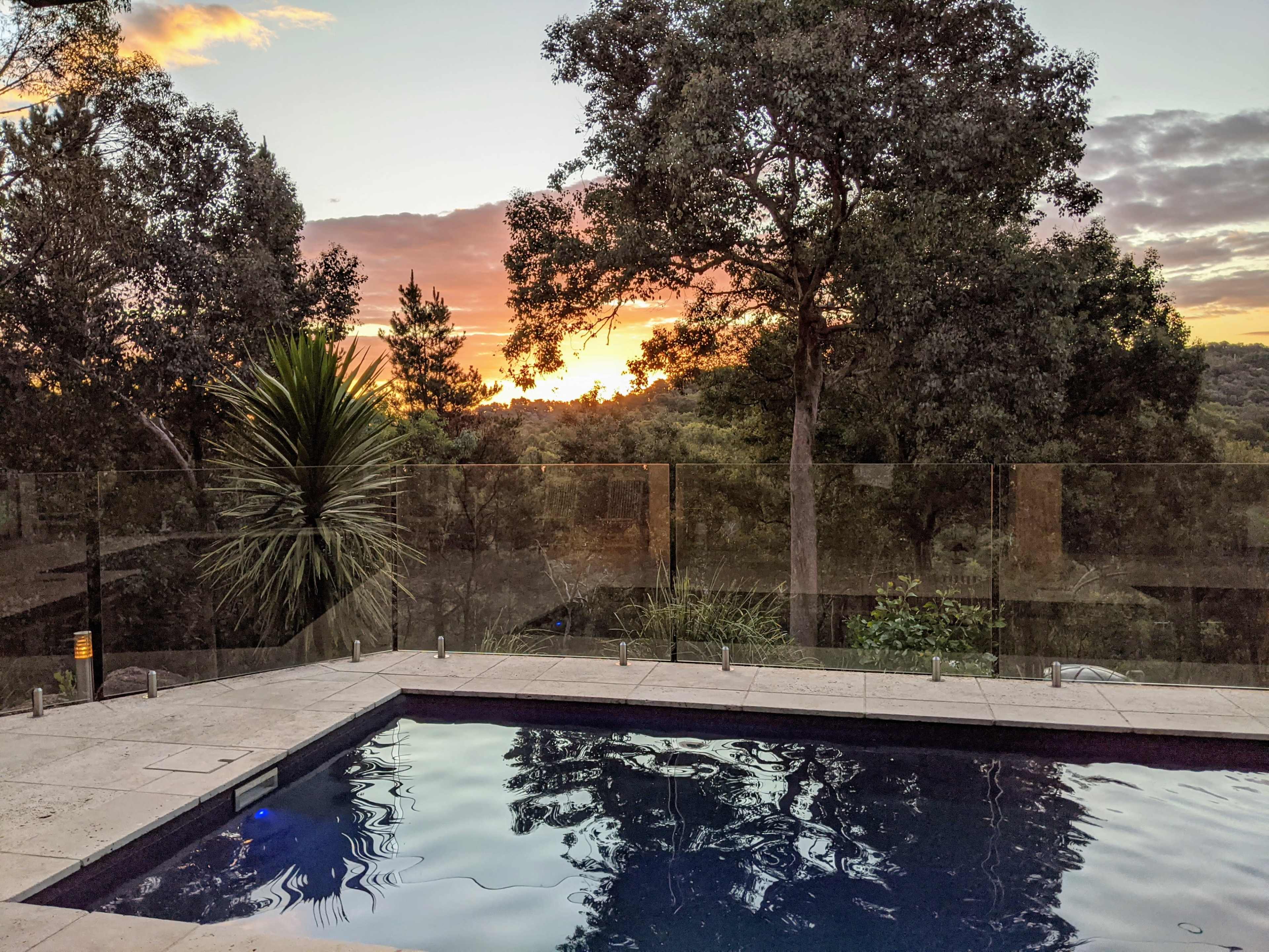 The image shows a pool surrounded by greenery with a view of a sunset over the horizon.