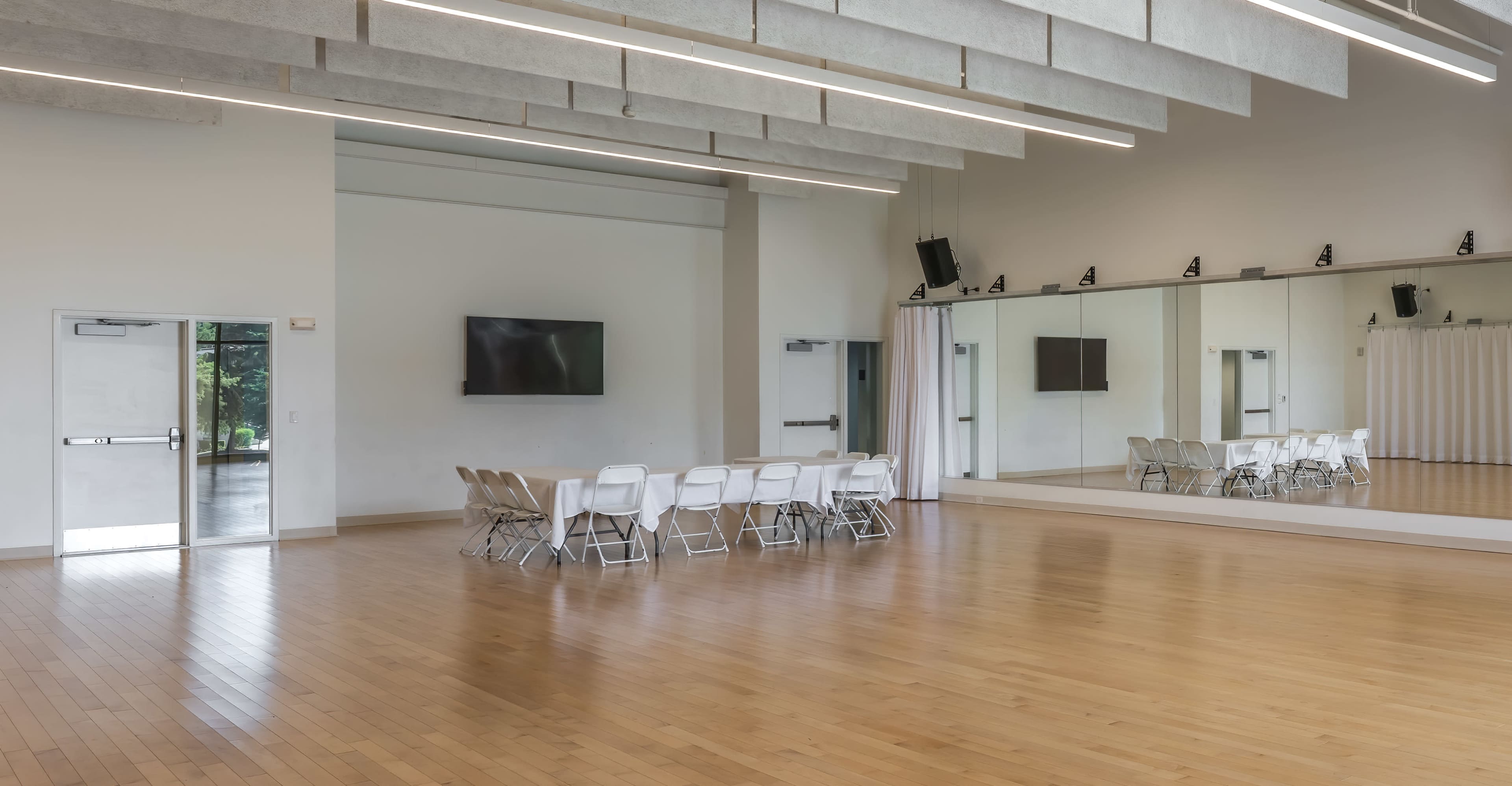 The image shows a spacious indoor room with wooden flooring, a mirrored wall, and a circular arrangement of white folding chairs.