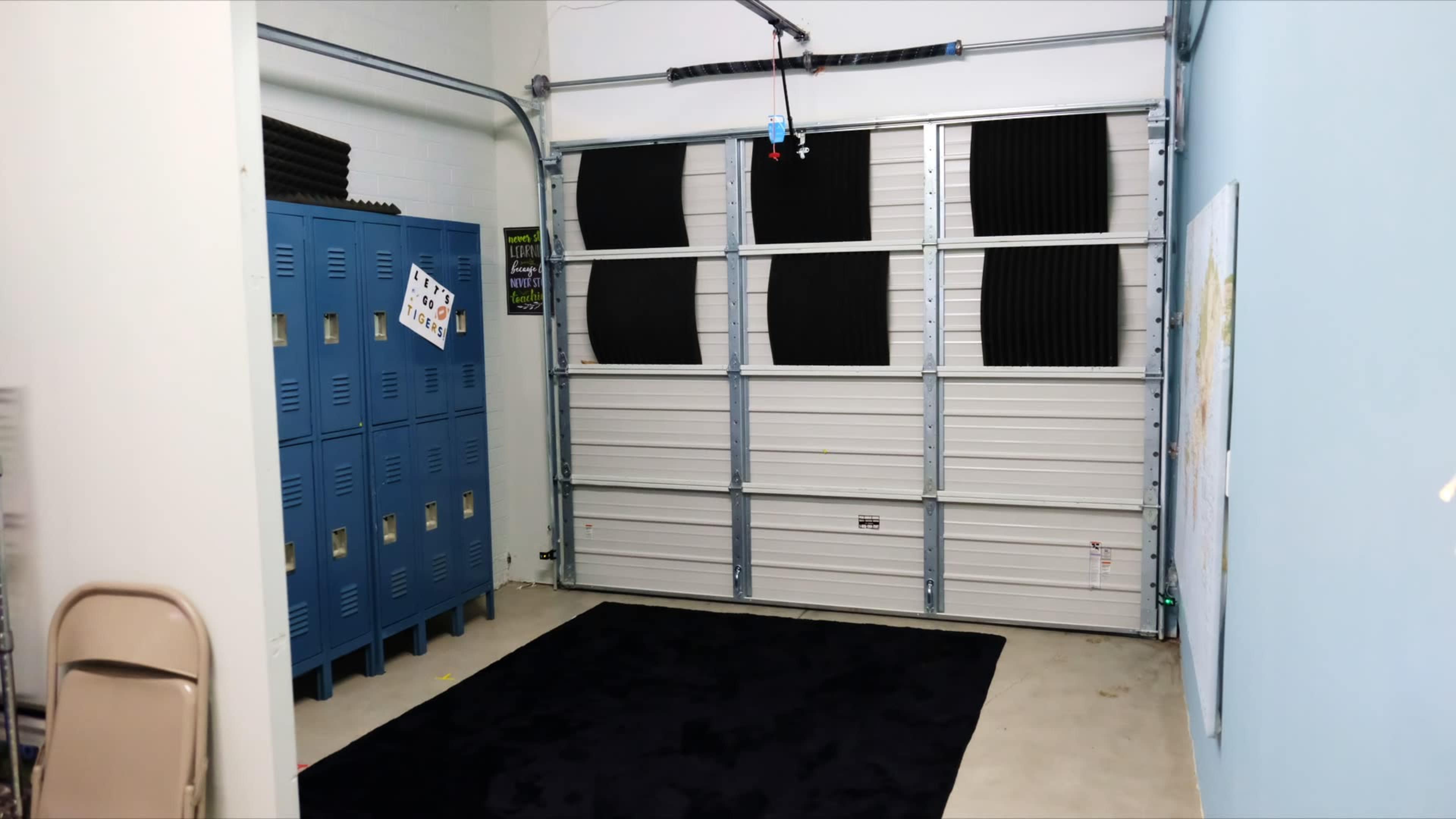 The image shows a garage interior featuring blue metal lockers on one side and a large garage door with window panels, complemented by a dark rug on the floor.