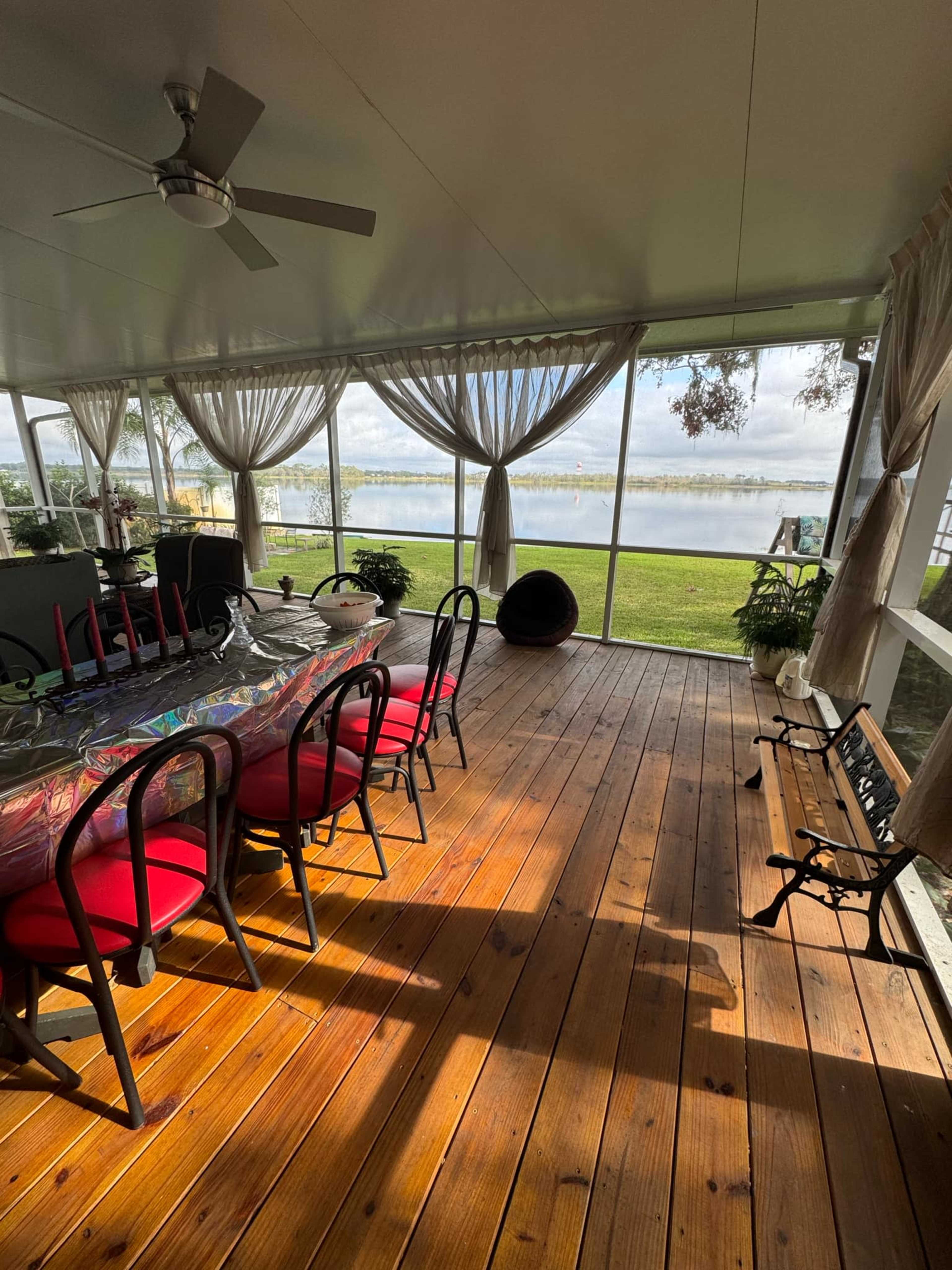 A screened porch features a wooden floor, a large dining table with red chairs, and a view overlooking a calm body of water.