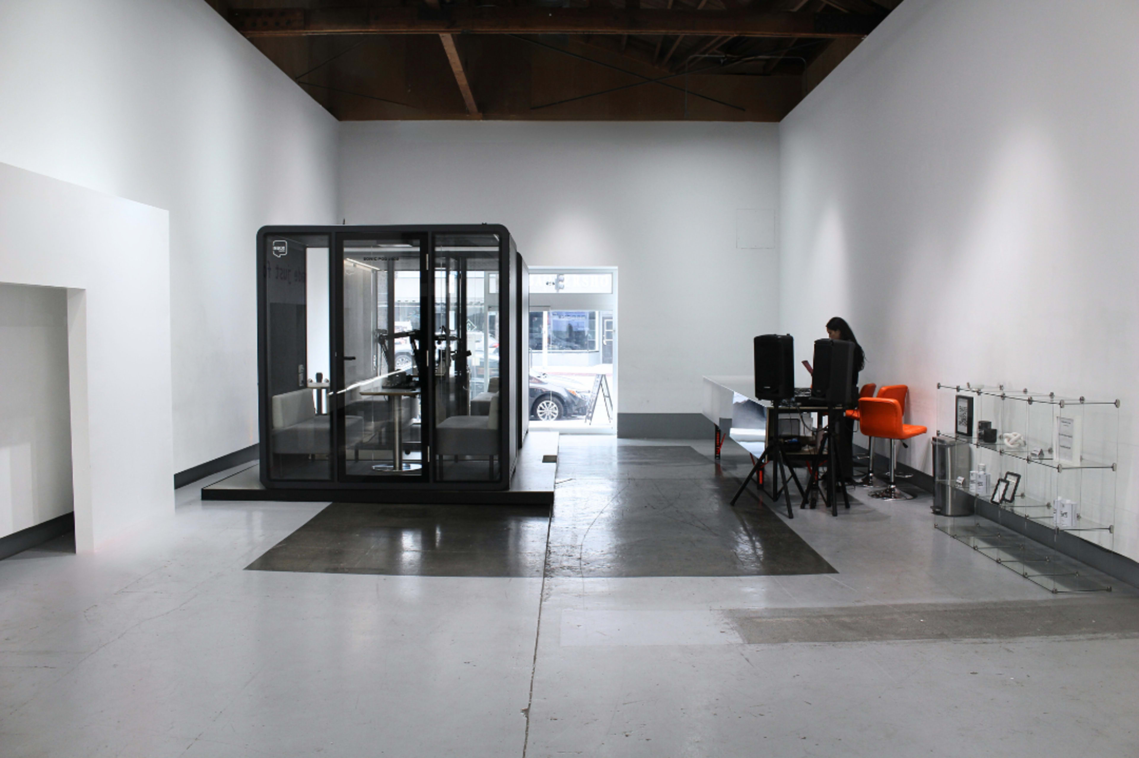 The image depicts a minimalist interior of a gallery or office space featuring a glass-enclosed booth, a desk with a chair, and a person working nearby.