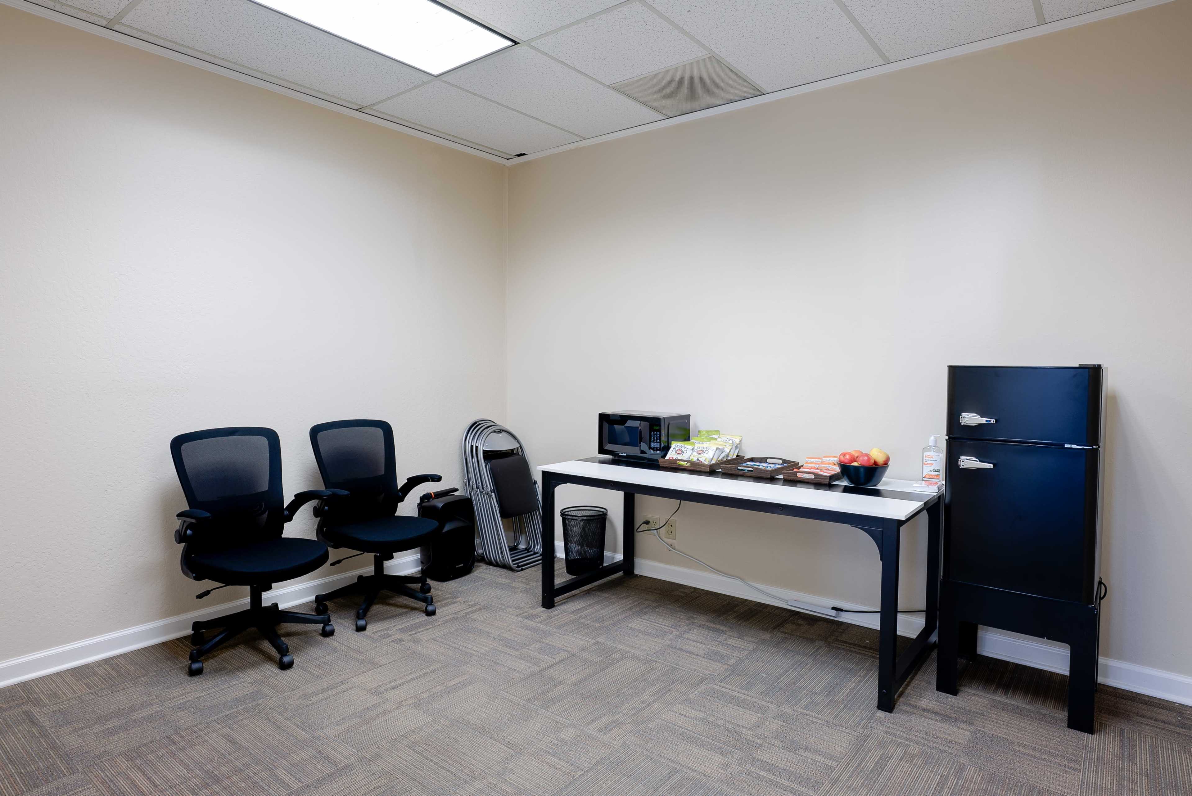 The image shows a small office space with two black chairs, a table displaying snacks and a microwave, and a black refrigerator along the wall.