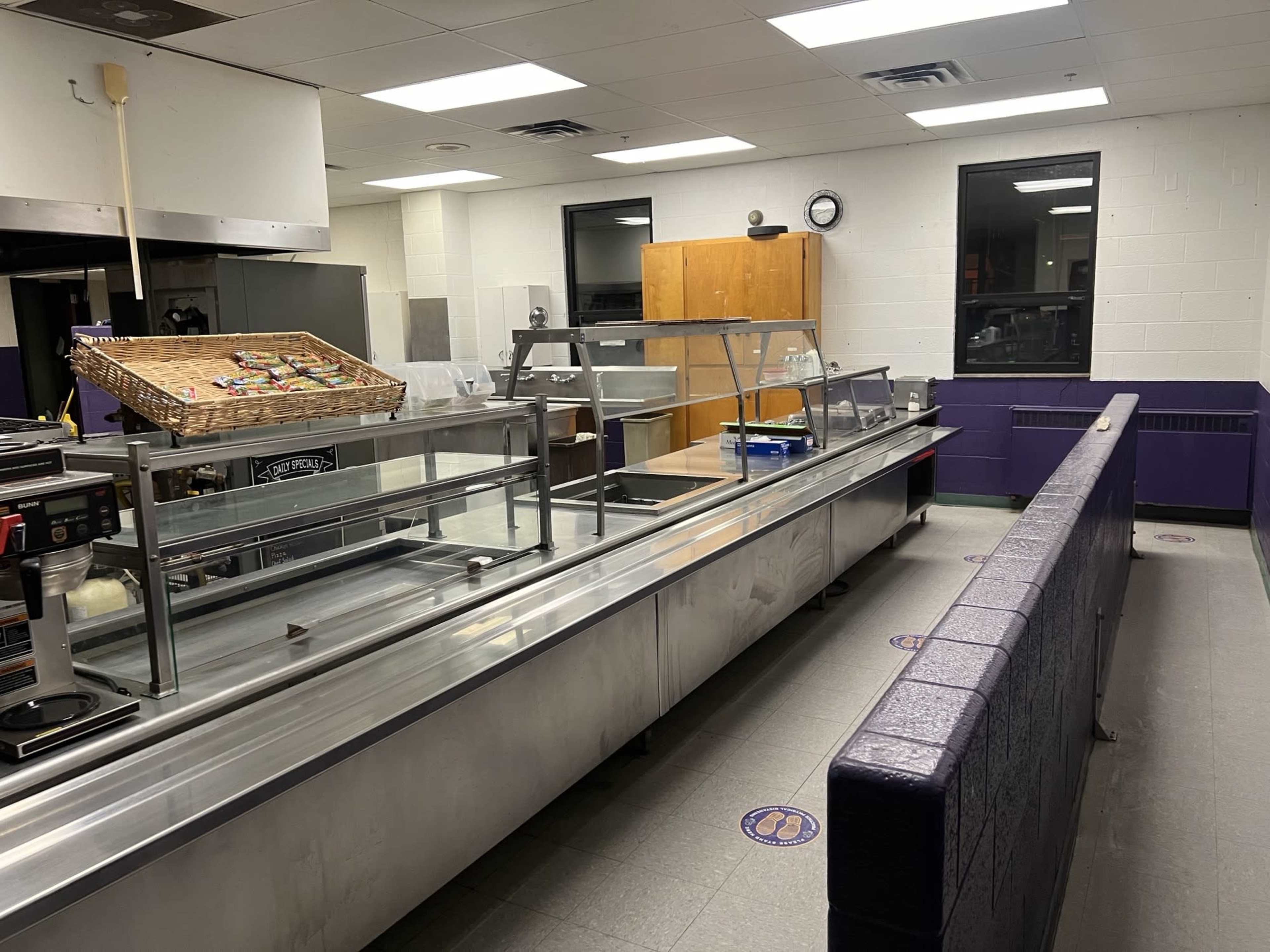 The image shows a cafeteria serving area with stainless steel food counters, a wooden storage unit, and a clock on the wall.