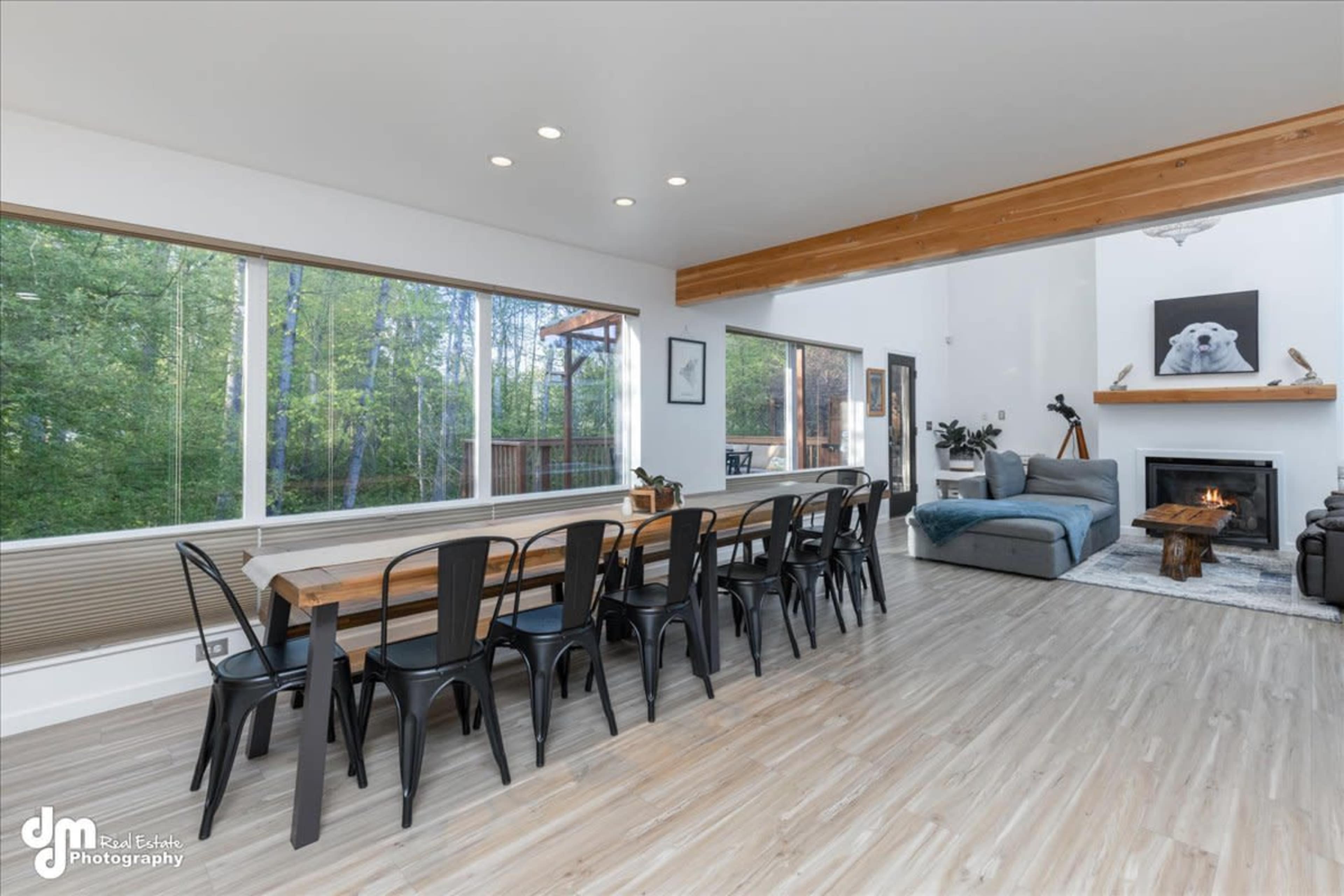 The image shows a modern dining area with a long wooden table surrounded by black chairs, adjacent to a cozy living space featuring a fireplace and large windows looking out into a green landscape.
