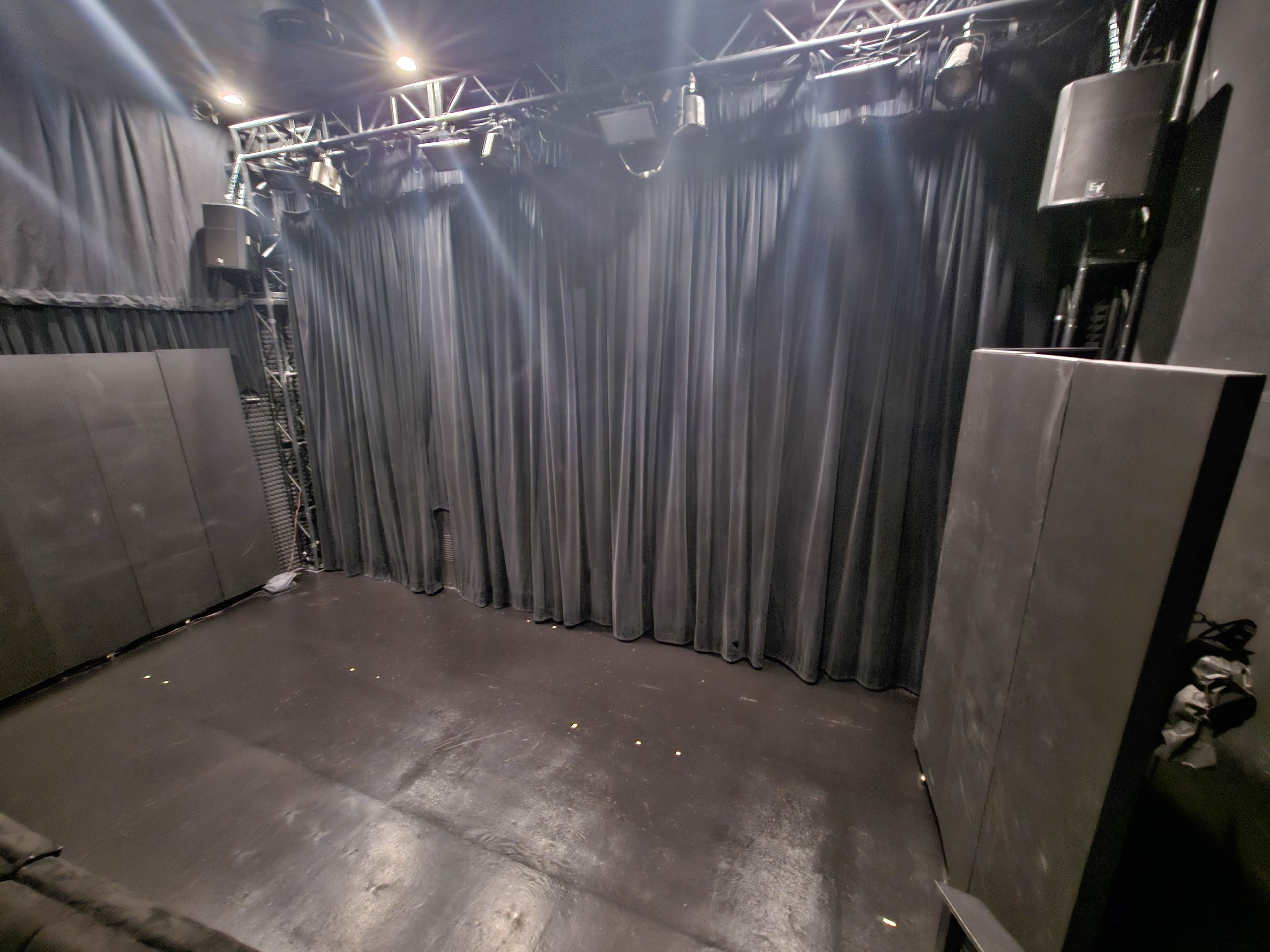 The image shows a darkened stage area with a black curtain backdrop, flanked by sound equipment and padded panels.