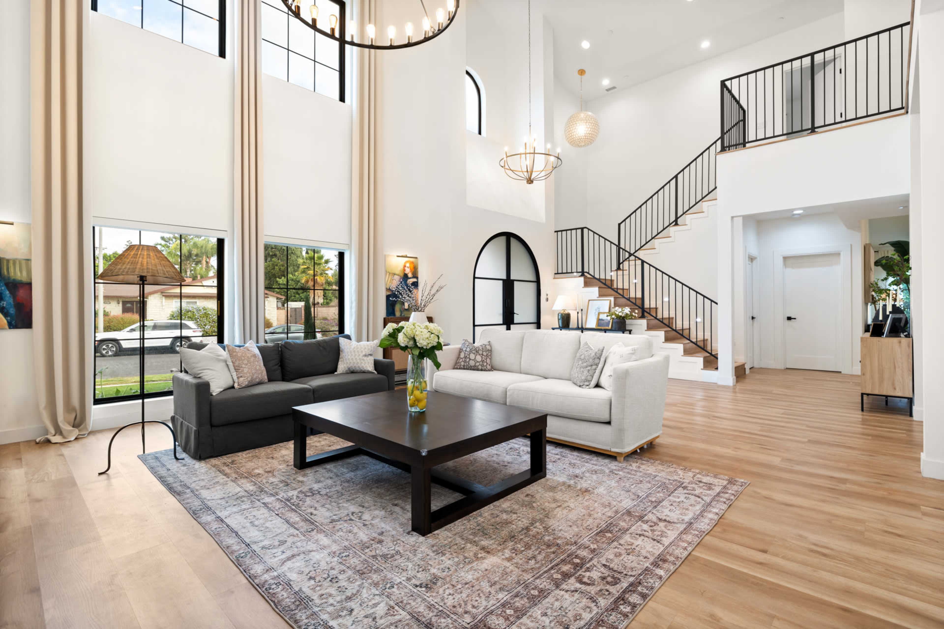 The living room features a light-colored sofa, a dark coffee table, large windows with views of the outdoors, and a staircase leading to an upper level.