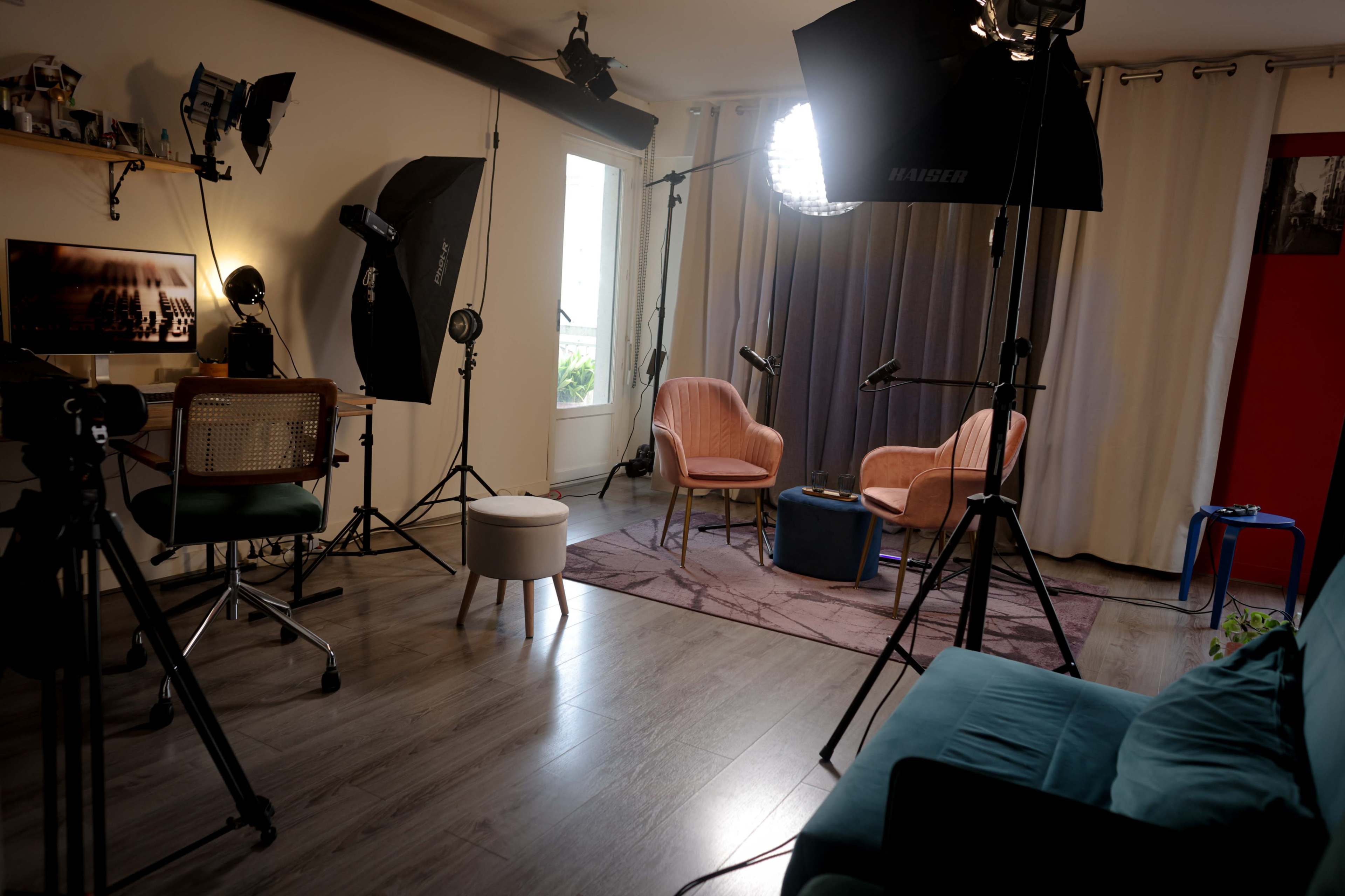 The image shows a well-lit studio space featuring two pink chairs, a small white ottoman, a blue table, and various lighting equipment arranged around the room.