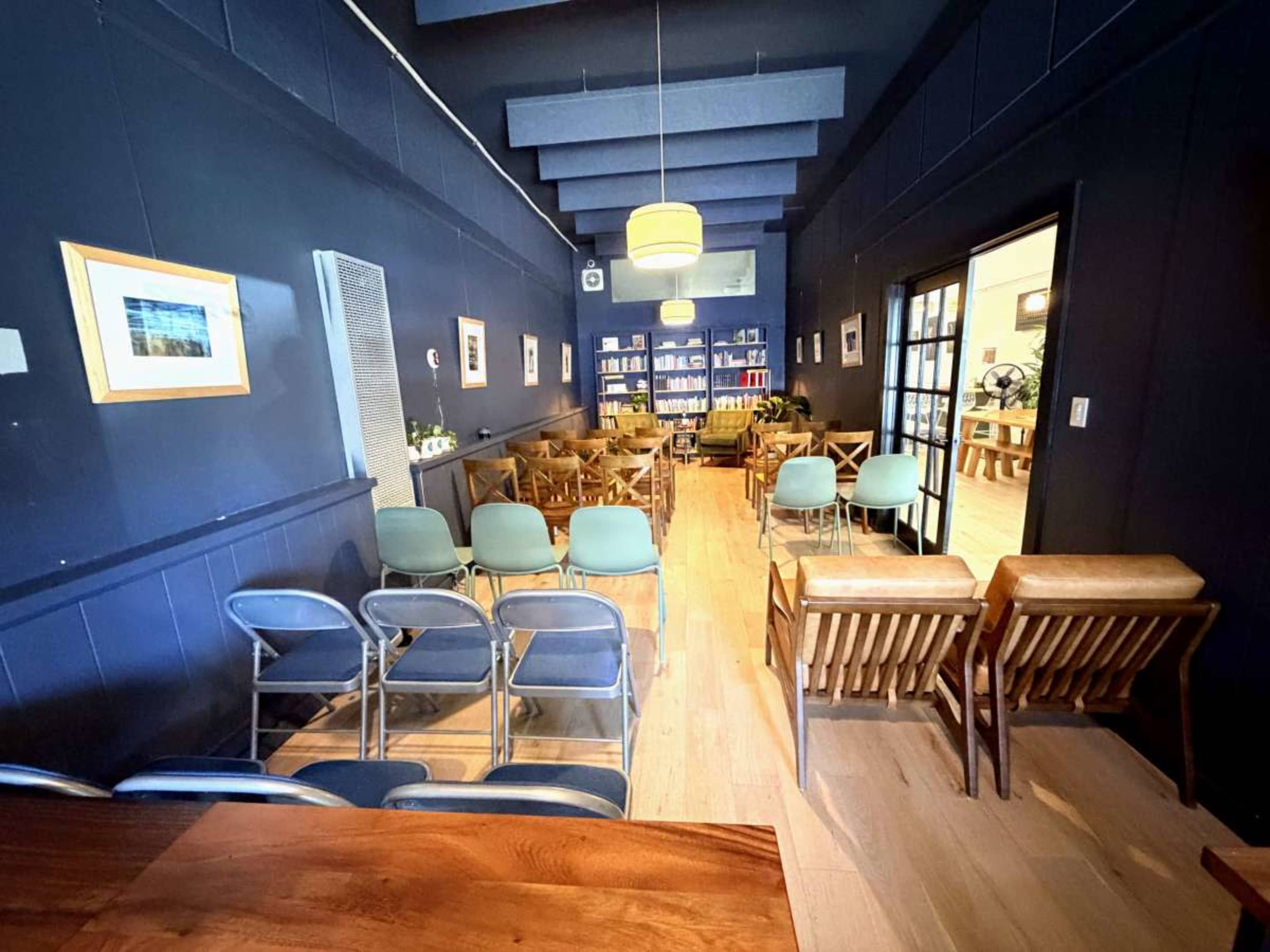The image shows a room with a dark blue wall, featuring multiple rows of chairs, a bookshelf in the back, and wooden elements in the furniture.