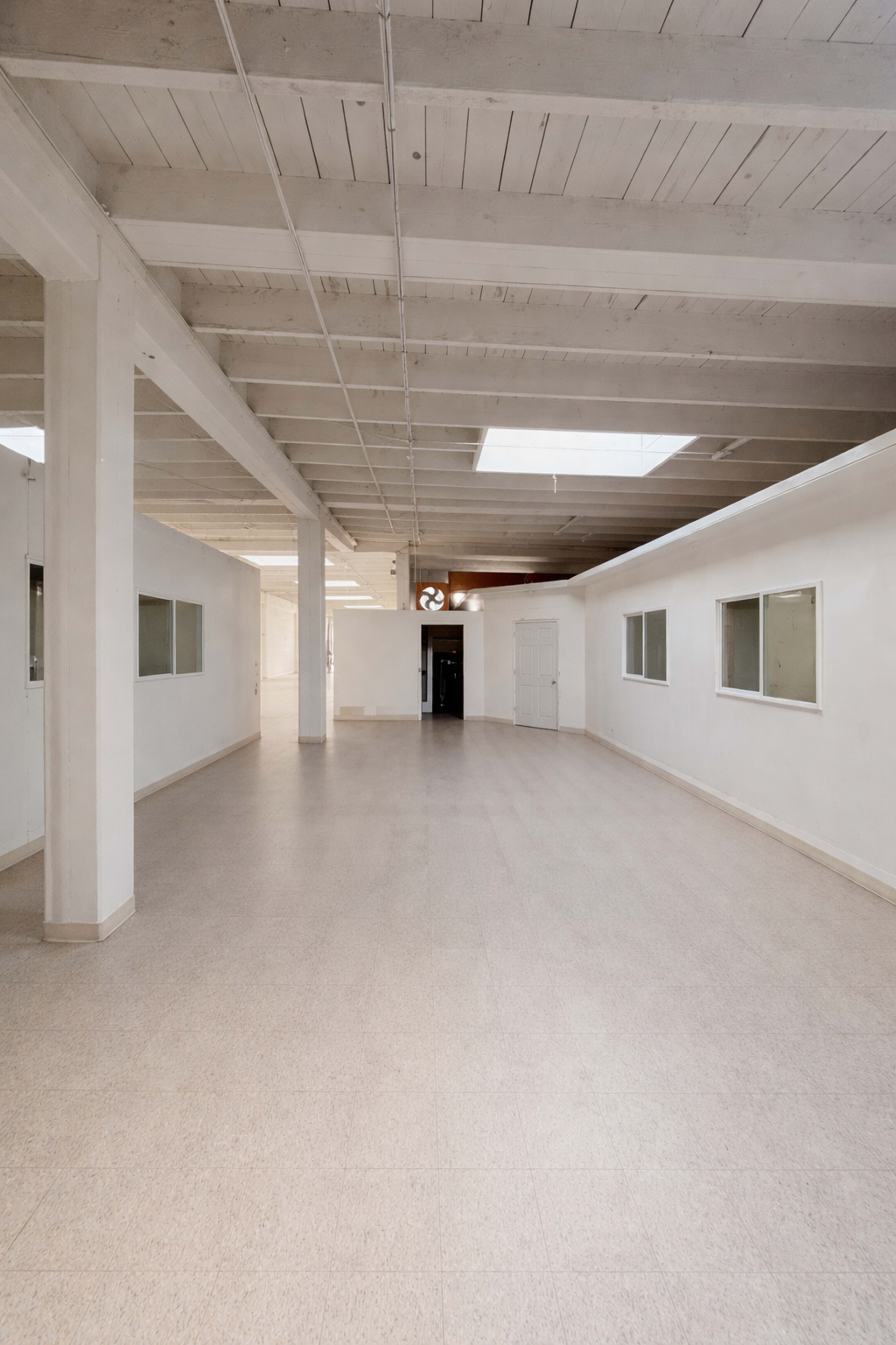 The image shows an empty, well-lit hallway with a high ceiling, featuring large windows on either side and a door at the far end.