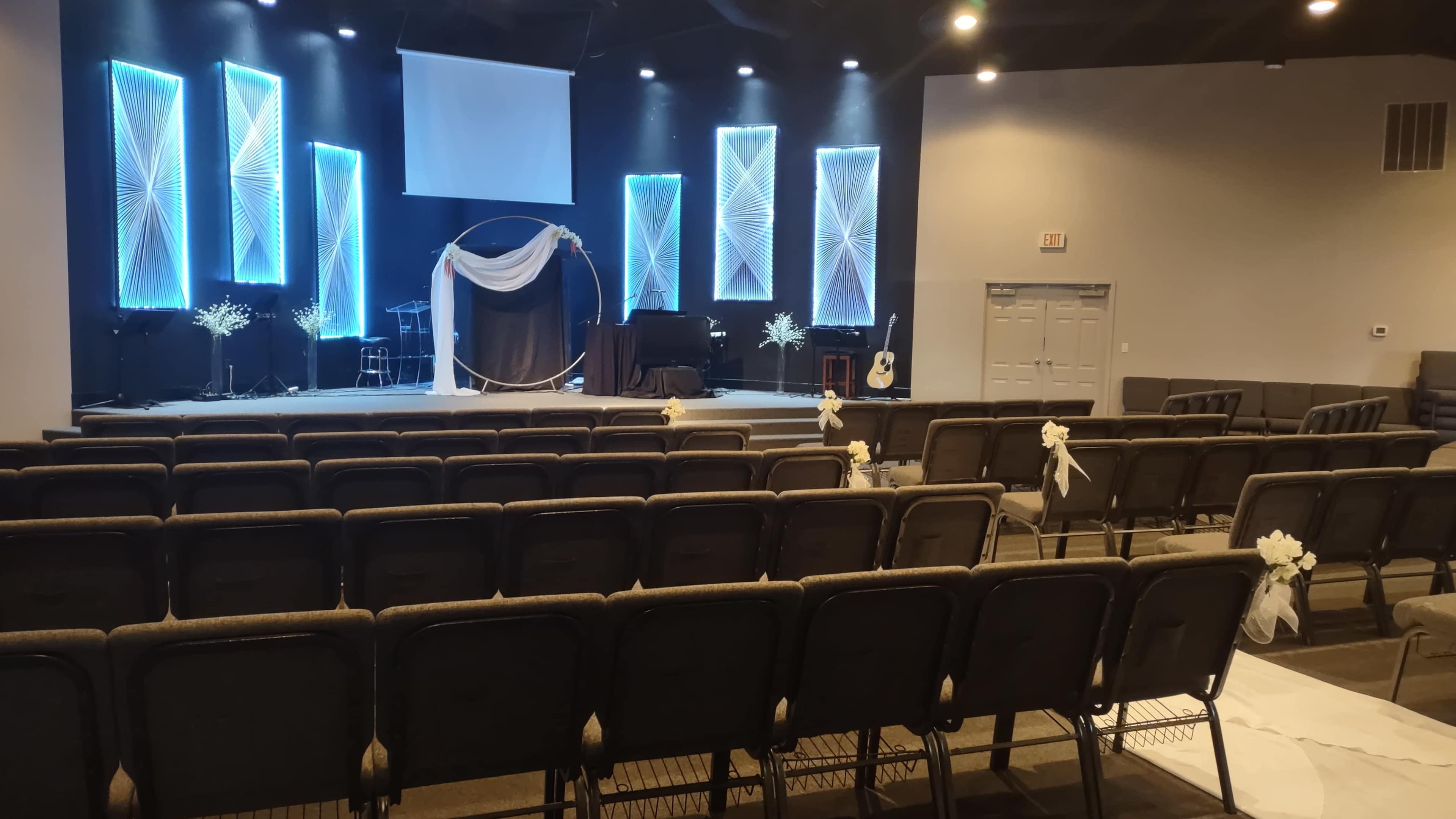 An empty auditorium set up for an event, featuring a stage with lighting fixtures, a backdrop draped in fabric, and rows of chairs arranged in front of it.