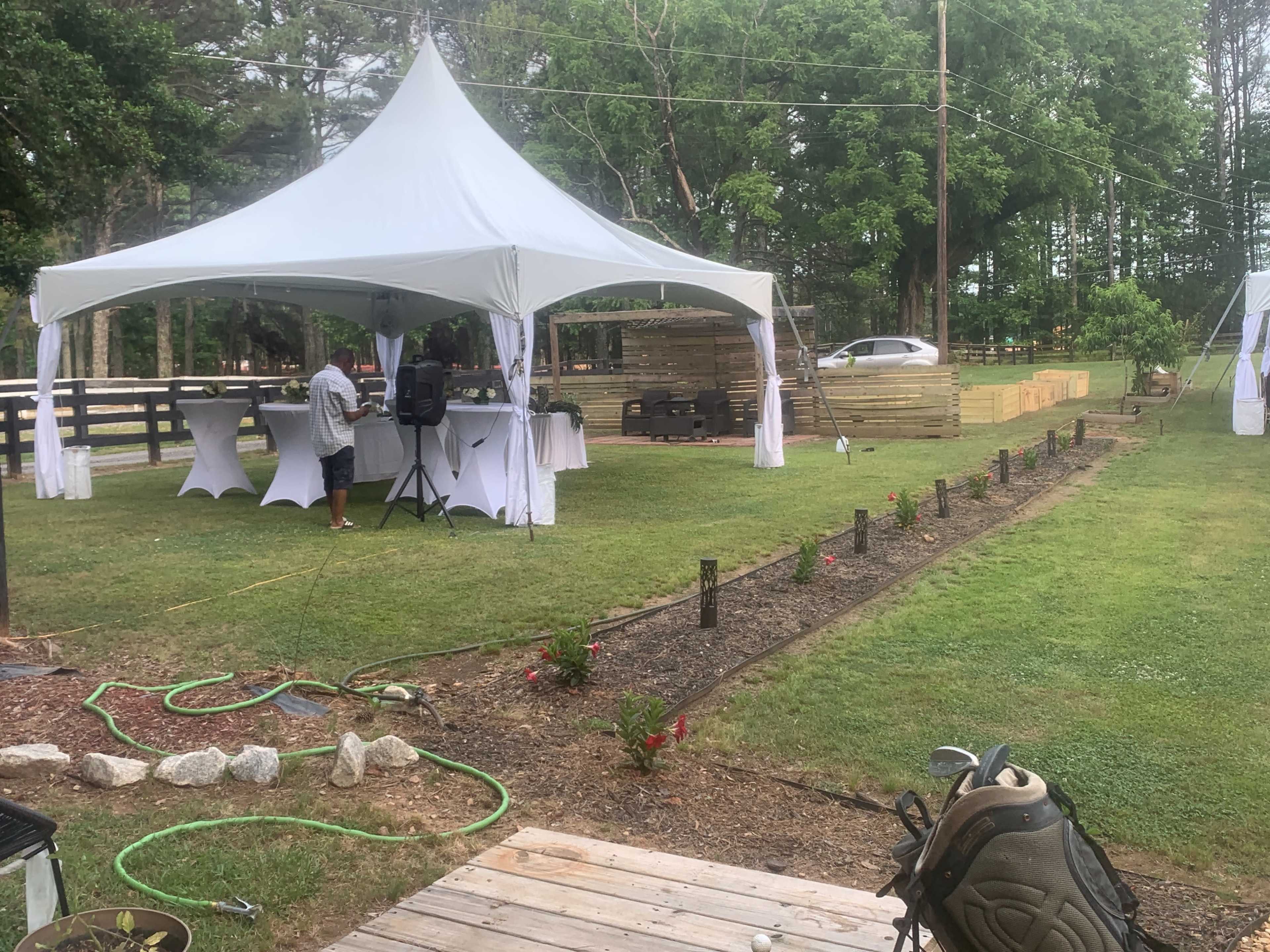 A white tent is set up on a grassy area with tables underneath, while a person prepares for an event in the background, and a pathway lined with flowers leads through the yard.