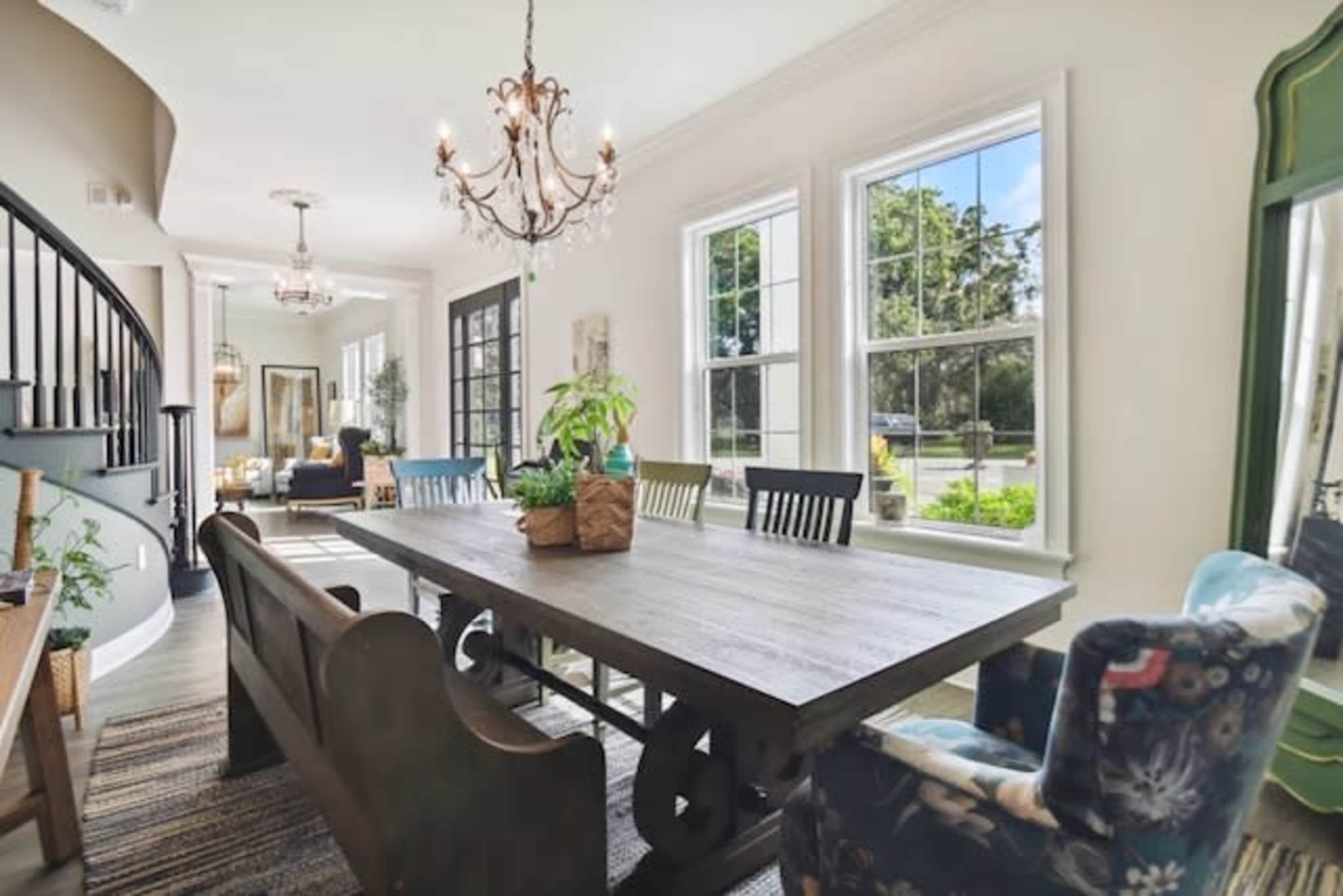 The image shows a bright dining area with a large wooden table surrounded by various chairs, a chandelier overhead, and large windows letting in natural light.