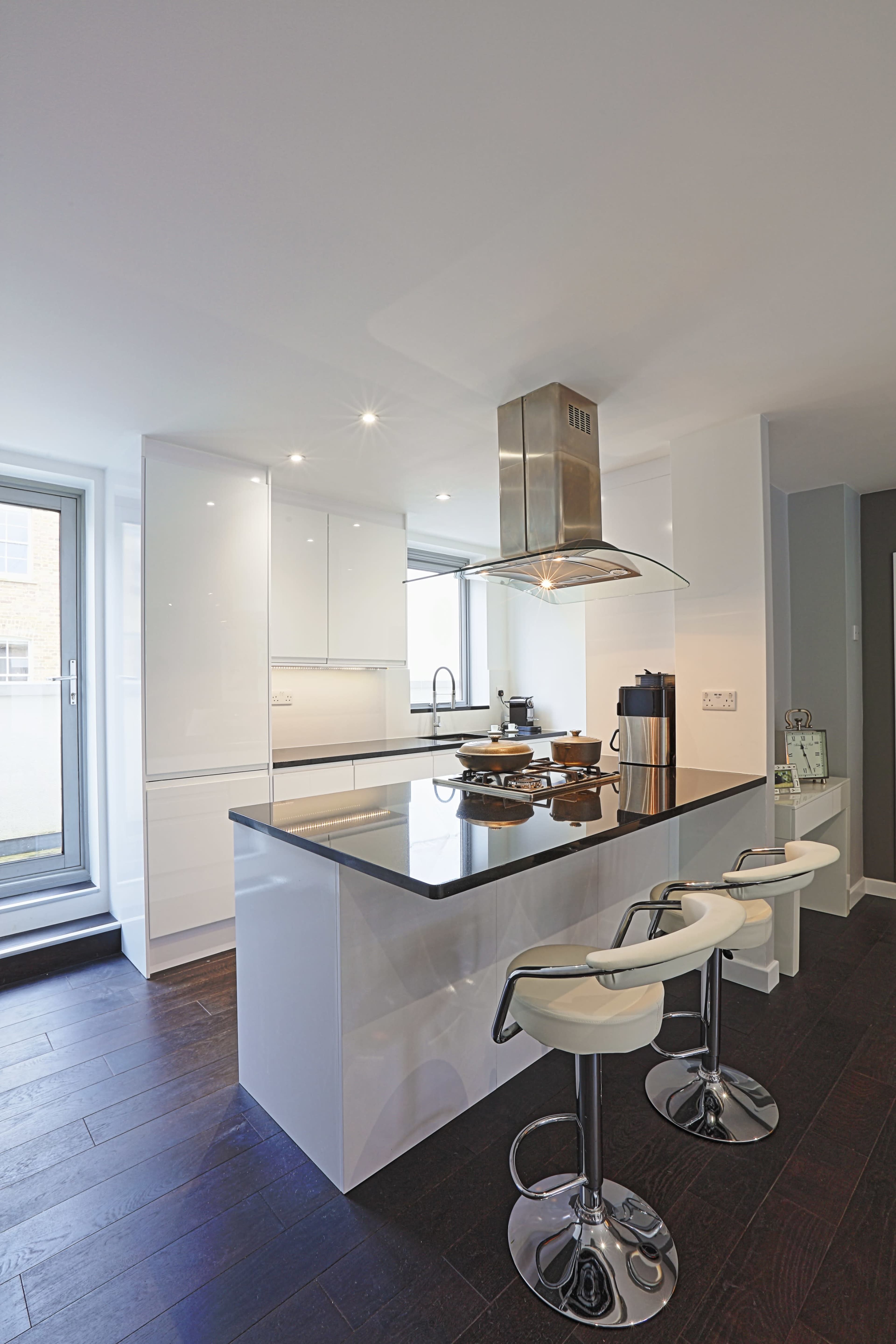 A modern kitchen features a central island with bar stools, white cabinets, and a stainless steel hood above the stove.