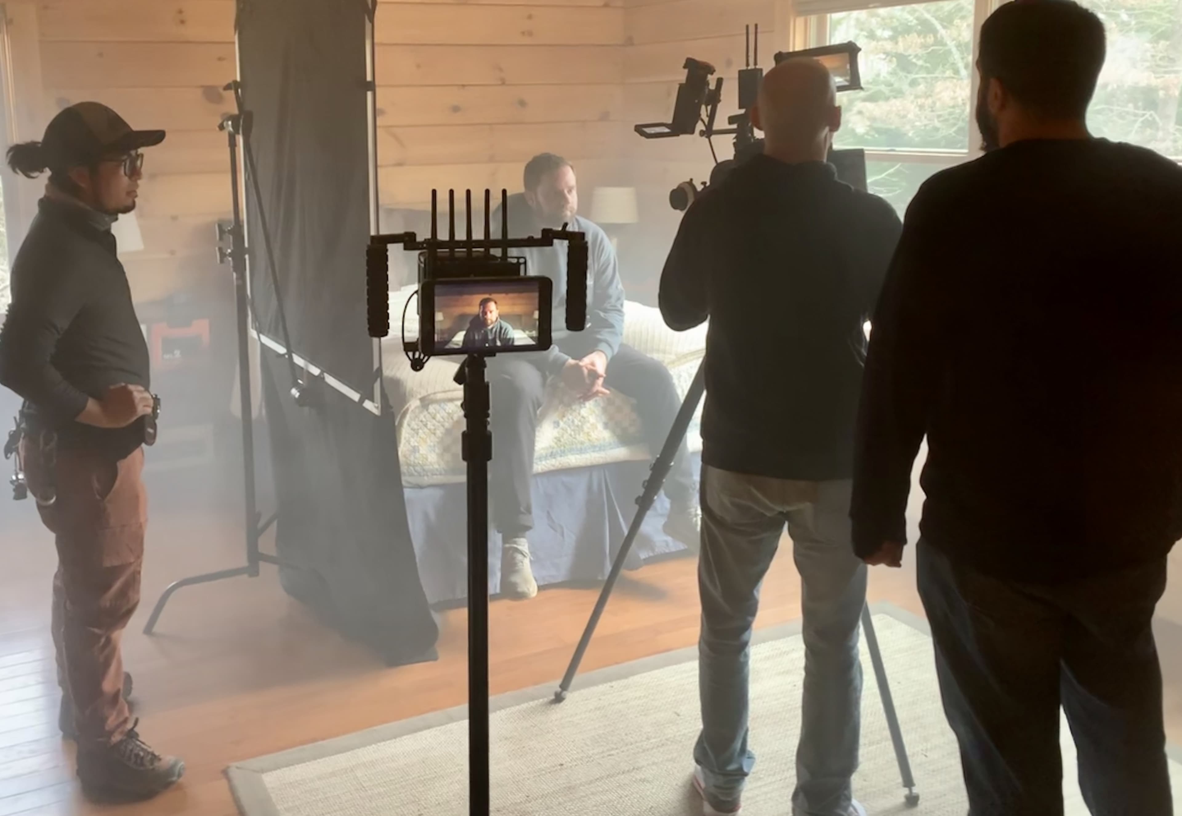 A film crew sets up a shot in a wooden room with fog, focusing on a seated subject while using multiple cameras and lights.