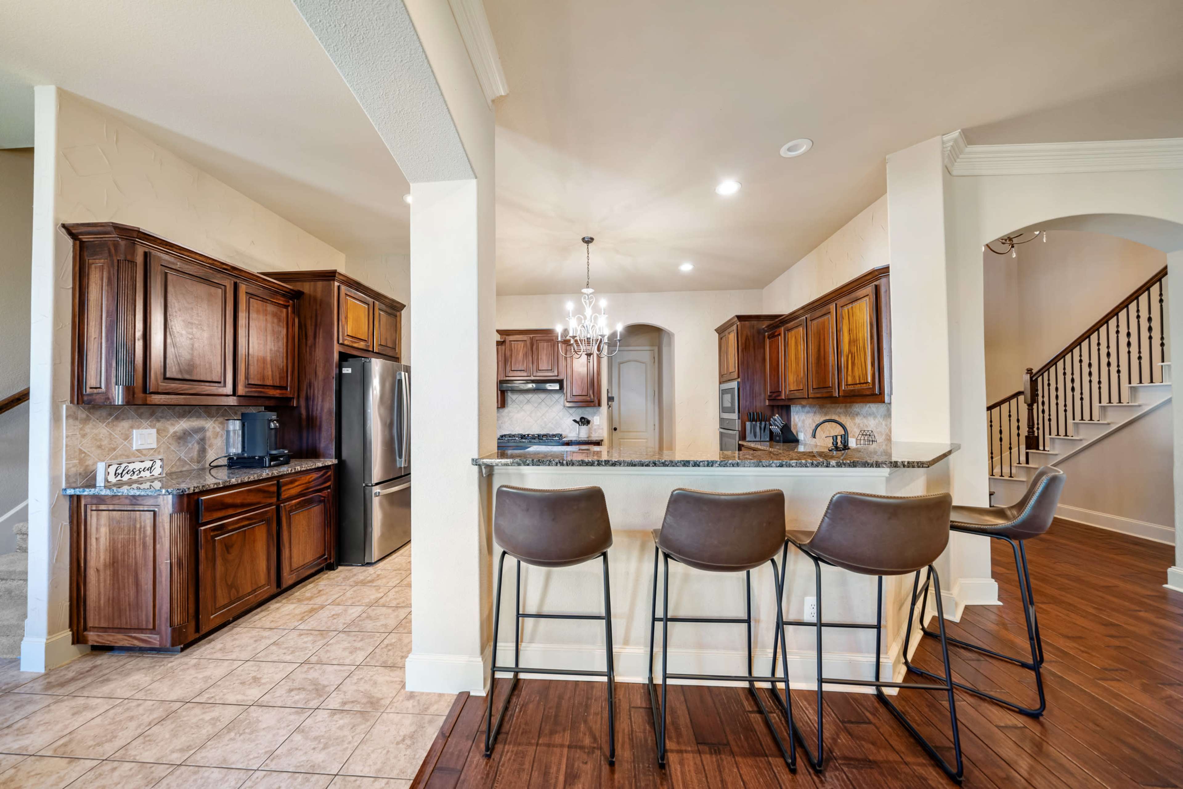 The image shows a modern kitchen with wooden cabinets, a granite countertop, and a breakfast bar with four stools.